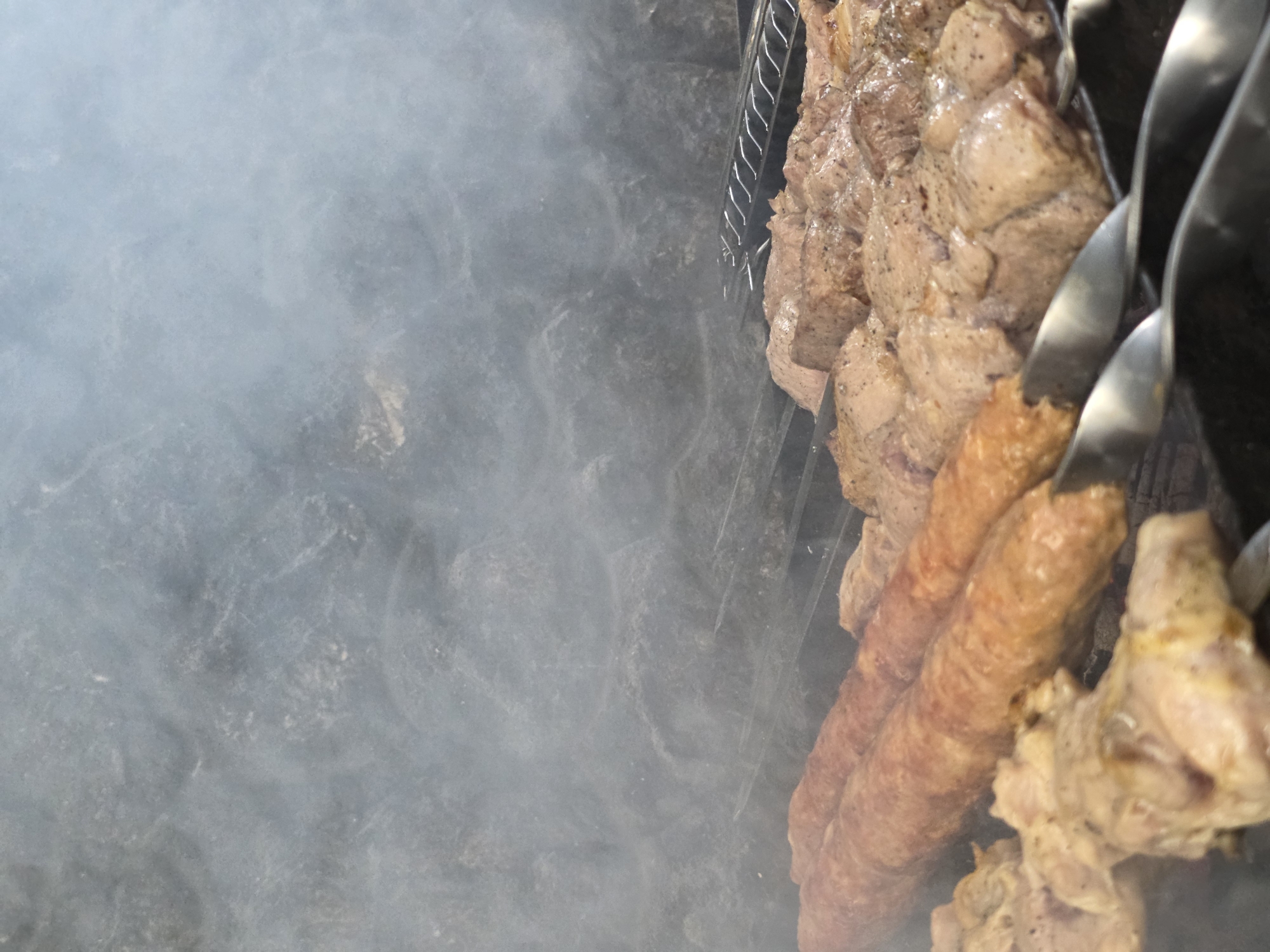 The media file shows a close-up, low-angle shot of meat cooking on skewers over a grill, partially obscured by smoke. The meat appears to be a mix of cubed pieces and cylindrical sausages, seasoned with pepper and possibly other spices. Several metal skewers are visible, holding the meat and resting on the grill grate. The background is dominated by the hazy, grey smoke, creating a diffused effect on the dark, textured surface beneath the grill. The scene is likely outdoors, given the open-air cooking and the presence of smoke. No people are visible in the frame. The lighting suggests it could be daytime, but the exact time is not discernible due to the smoke and angle. The location is identified as Pidhorodne, Ukraine. No text is visible in the image.