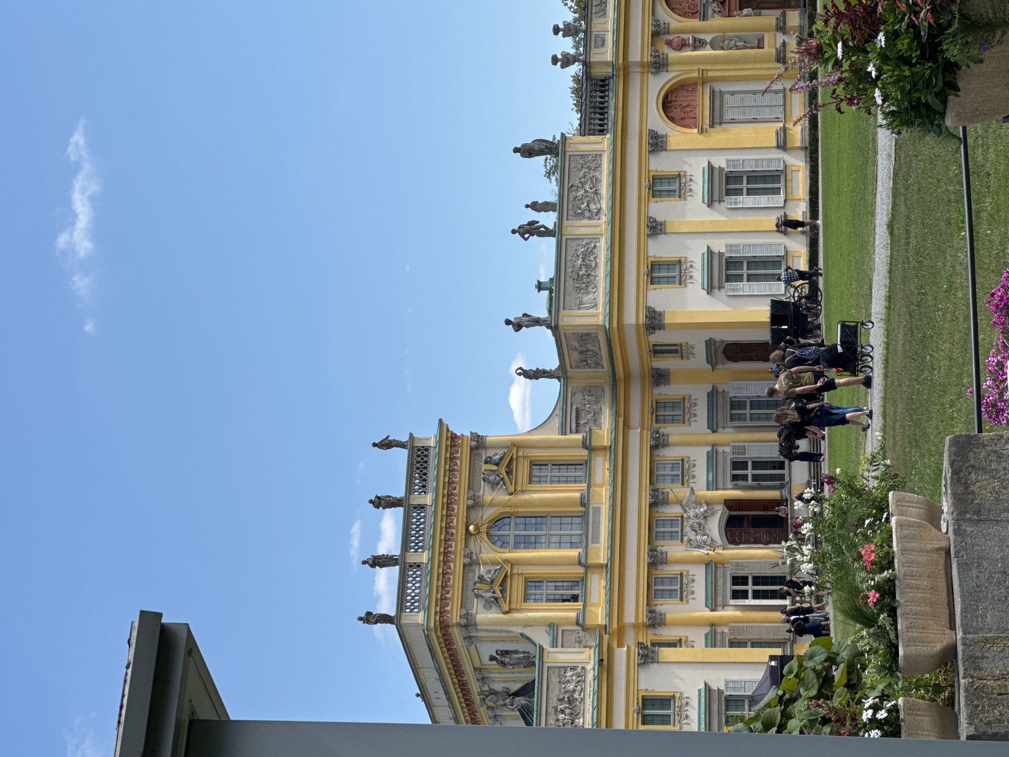The image shows the Wilanów Palace, a historical royal palace located in Warsaw, Poland. The palace is a grand yellow Baroque building with ornate architectural details, including columns, sculptures on the roofline, and decorative friezes. The building is set against a clear blue sky with a few scattered clouds. In front of the palace, there is a well-maintained green lawn with several planters filled with flowers. A group of people are visible on the lawn, some walking and others standing. There is also a horse-drawn carriage in the middle ground. The foreground is partially obscured by a gray structure on the left and planters with flowers on the right. The overall scene depicts a sunny day at a prominent historical landmark.
