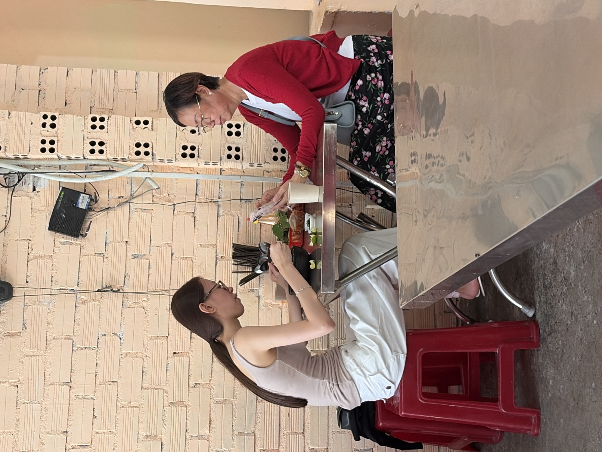 Two women are seated at a table in what appears to be an outdoor eatery in Nha Trang, Vietnam. The woman on the left, with long dark hair pulled back in a ponytail, wears a light-colored tank top and loose white pants. She is looking down at a bowl of food and using chopsticks. She is seated on a red plastic stool.
The woman on the right has short dark hair and wears glasses, a white t-shirt, and a red cardigan. She is holding a smartphone in her left hand and looking at it. She is wearing a patterned skirt. She has a light blue crossbody bag.
On the table between them are bowls of food, chopsticks, a cup, and what appears to be a small condiment dish. The background shows a brick wall with exposed wiring and electrical conduit, and part of a light-colored wall. The table in the foreground has a reflective surface. The overall environment suggests a casual dining setting.