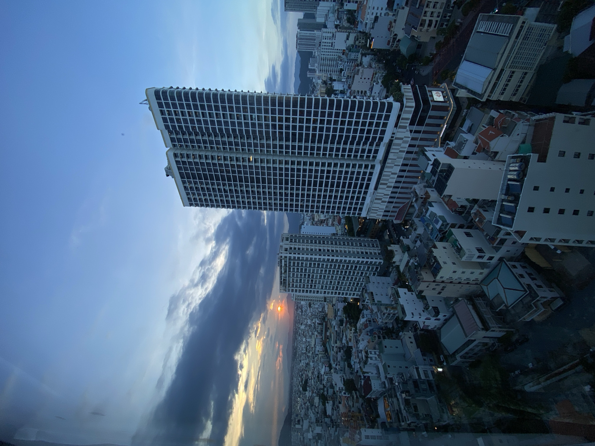 This is a high-angle, wide shot of a cityscape at dusk, viewed through a window, likely from a tall building in Nha Trang, Vietnam. The foreground is dominated by the reflection of the photographer and the window frame, which distorts the scene. In the middle ground, two prominent, modern high-rise buildings stand out. The taller one on the right has a white facade with a grid of dark windows. The building to its left is also a high-rise, appearing slightly shorter and with a similar architectural style.
Beyond these buildings, the city stretches out, a dense collection of smaller residential and commercial buildings with a variety of roof colors, including red and blue. The sky is a mix of deep blue and grey clouds, with the sun setting in the distance, casting a warm orange glow through a break in the clouds. The silhouette of mountains is visible on the horizon, indicating the city's proximity to natural topography. The streetlights and building lights are beginning to illuminate, suggesting the transition from day to night. The overall impression is one of an urban environment at the end of the day, with a blend of modern architecture and more traditional city structures.