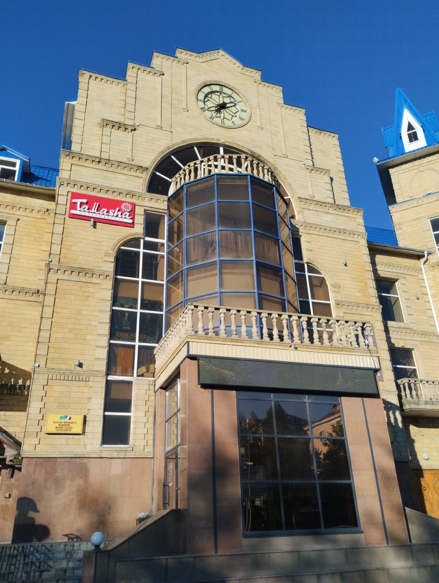 The image shows the facade of a building with a clock on top, a blue turret, and a red sign that says "Tallasha". The building is made of beige bricks and has large windows. There is a yellow sign with text in Kazakh and Russian on the left side of the building. The building appears to be a commercial establishment, possibly a supermarket, given the sign. The scene is set against a clear blue sky. The location of the building in Kostanay, Kazakhstan, is implied by the presence of signs with text in Kazakh.