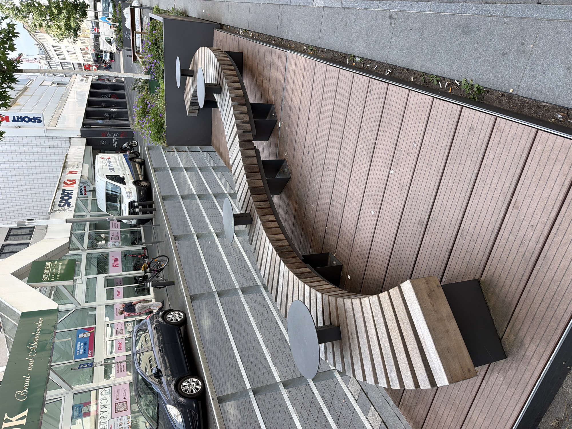 The image depicts an outdoor seating area on a sidewalk in Bochum, Germany, situated in front of storefronts. A curved, wooden slatted bench with integrated small round tables is the focal point, placed on a brown wooden deck. Behind the bench, a metal railing separates the seating area from the street, where a dark gray car is parked. Storefronts with large glass windows and signage are visible in the background. One sign reads "SCHODROK BRAUT- und ABENDMODEN," indicating a formal wear shop. Another sign shows "SPORT K Koch." A white delivery van is parked in front of one of the businesses. The environment appears to be a commercial district with urban elements.