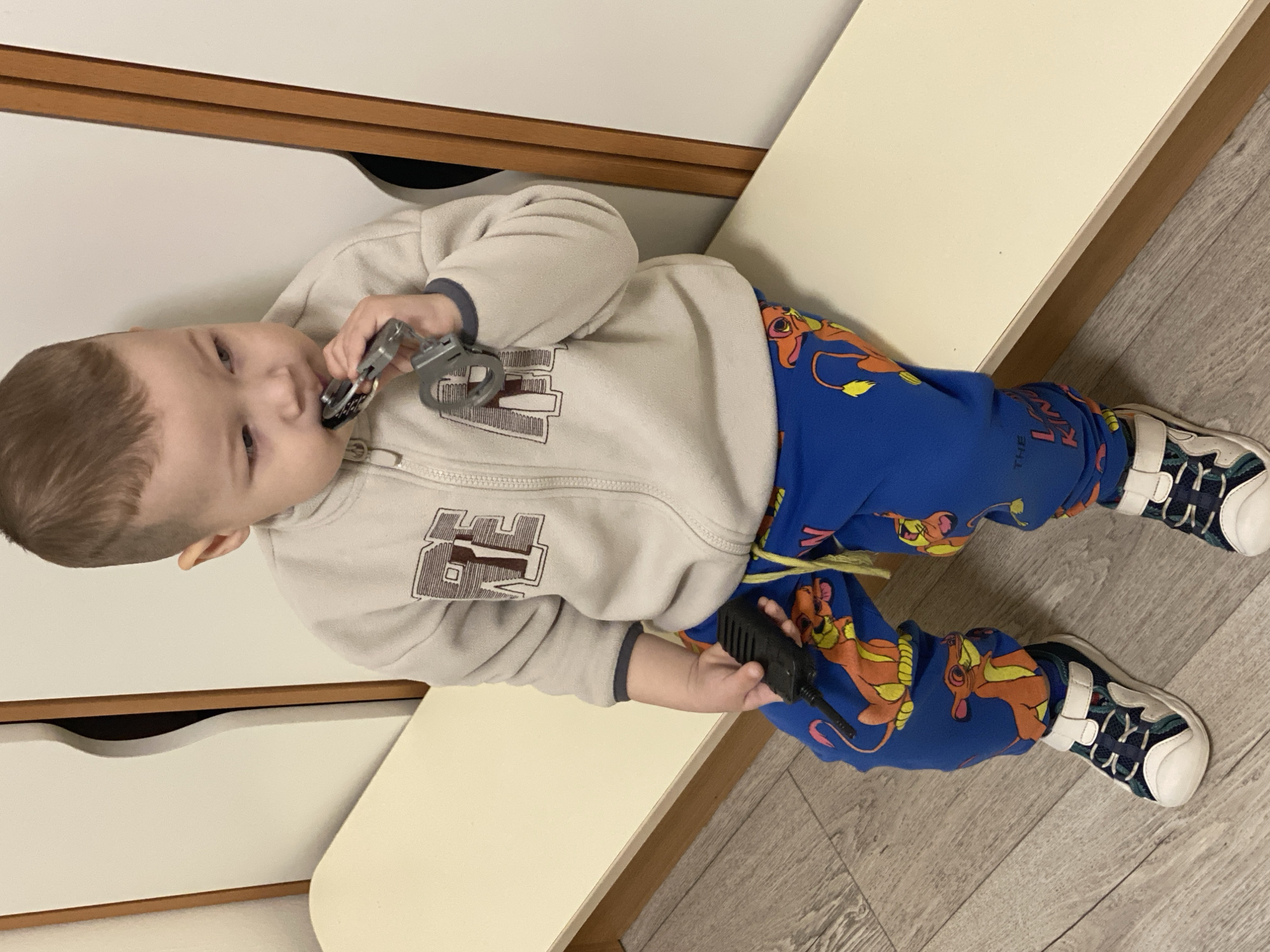 A young child is positioned at an angle, lying on a beige surface that appears to be part of furniture, possibly a bench or a low table. The setting seems to be indoors, likely a house or apartment. The child is dressed in blue pants patterned with images from The Lion King and a light-colored zip-up hooded sweatshirt with a brown graphic on the chest.
The child is holding two items. In their left hand, near their mouth, they are holding a pair of toy handcuffs. In their right hand, they are holding a black walkie-talkie.
The floor is a light-colored woodgrain. Adjacent to the beige surface is a white cabinet or storage unit with recessed drawer pulls.
There is no evidence indicating the scene is in Sochi, Russia.