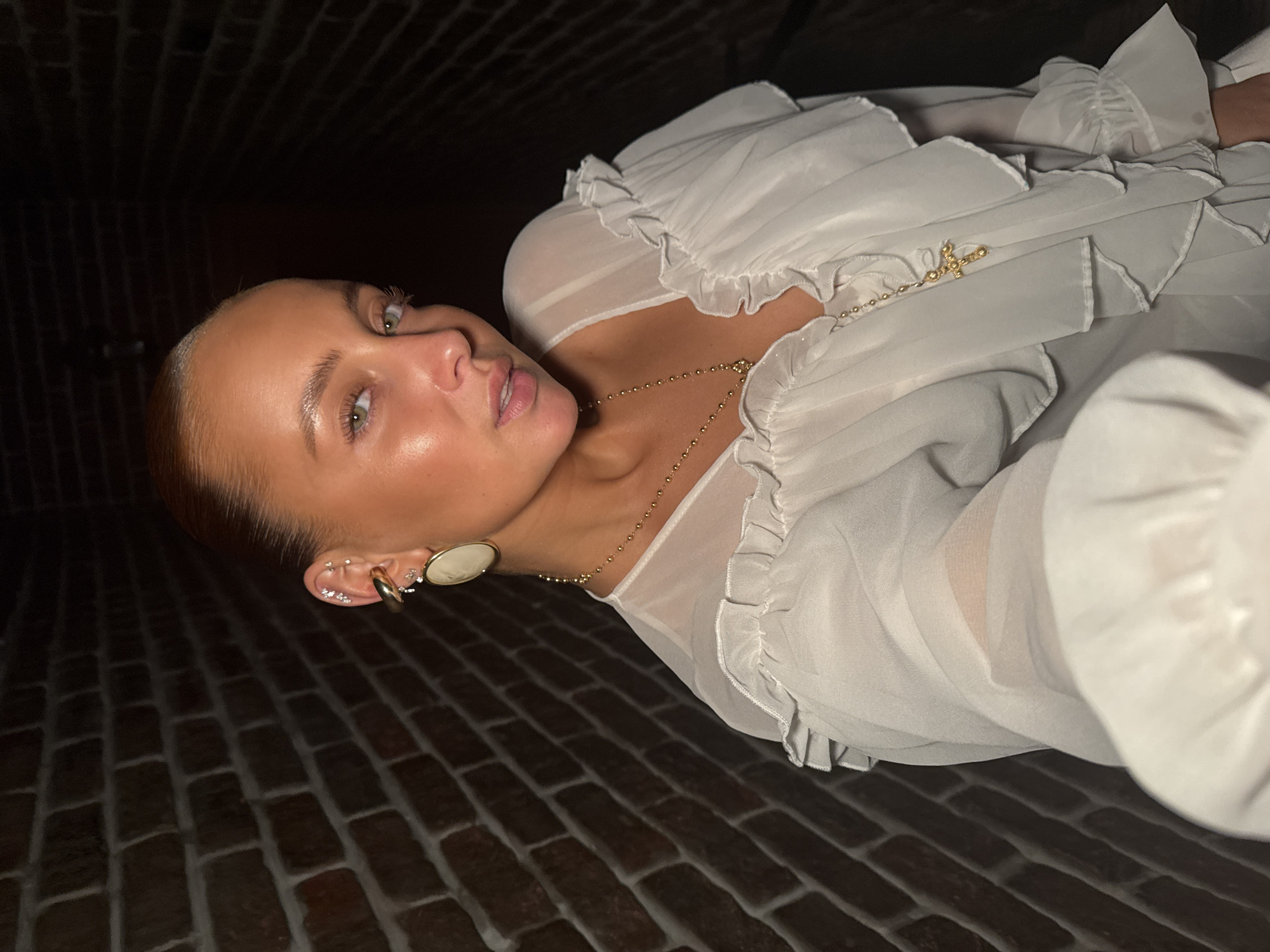 A woman is taking a selfie in a dimly lit brick hallway. She has her hair pulled back and is wearing a white ruffled blouse and gold jewelry, including earrings and a necklace with a cross pendant. The brick wall behind her is dark and textured. The image is cropped closely to the woman's face and upper body. The specific location in the city of Chaikovsky, Russia is not discernible from the provided image.