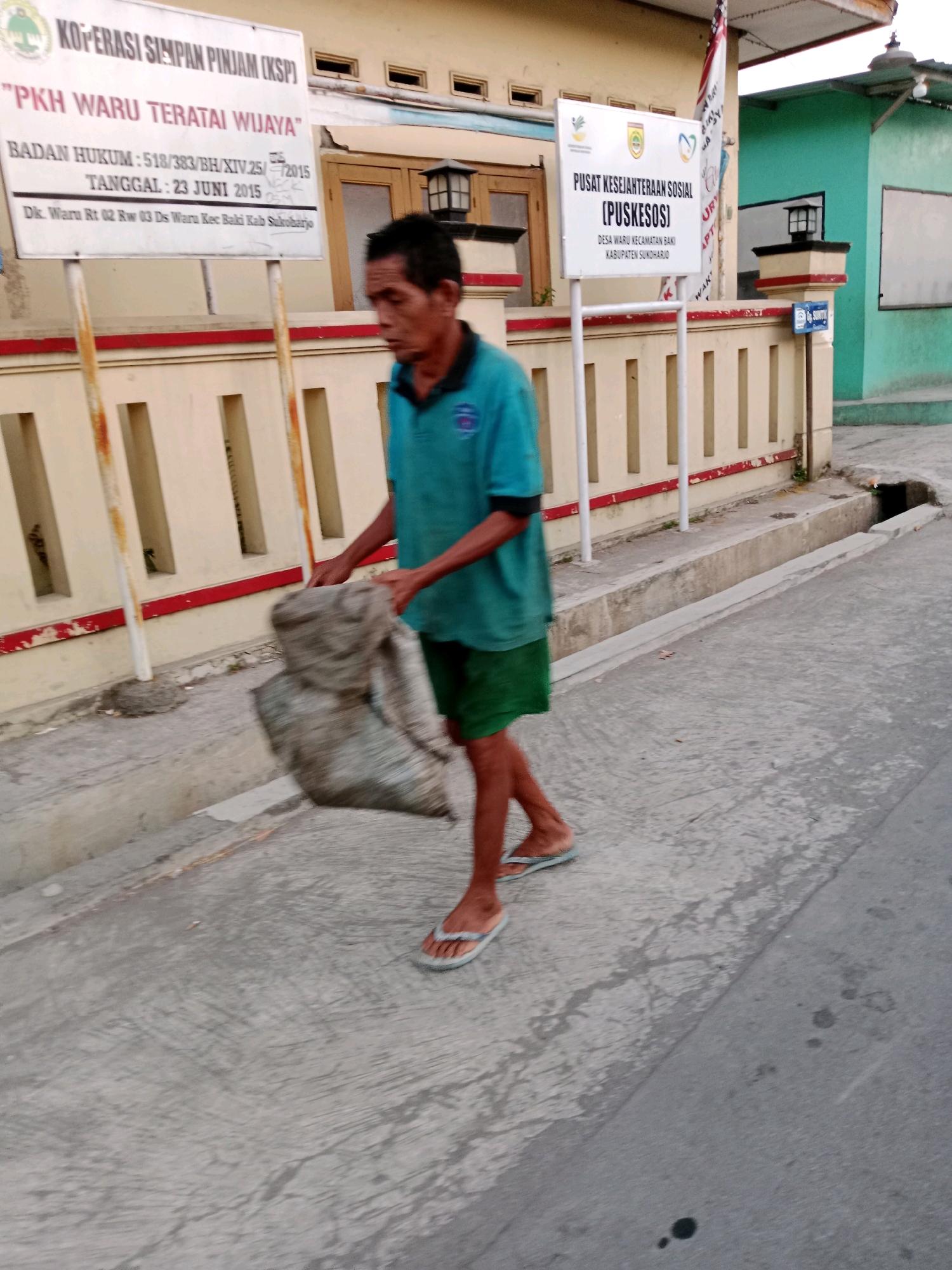 The image depicts a street scene in Waru, Indonesia. A man, wearing a teal t-shirt and green shorts, is walking on a concrete sidewalk carrying a large, gray, fabric-wrapped bundle in his arms. He is wearing flip-flops. To the left of the man, a white sign with black text is mounted on metal posts. The sign displays information about "KOPERASI SIMPAN PINJAM (KSP) 'PKH WARU TERATAI WIJAYA'" and lists administrative details like "BADAN HUKUM : 518/383/BH/XIV.25/2015" and "TANGGAL : 23 JUNI 2015". Further text on the sign indicates an address: "DK. Waru Rt 02 Rw 03 Ds Waru Kec. Baki Kab Sukoharjo". To the right of the man, another white sign, also on metal posts, reads "PUSAT KESEJAHTERAAN SOSIAL (PUKSOS)" and provides location details "DESA WARU KECAMATAN BAKI KABUPATEN SUKOHARJO". The buildings in the background are light yellow with brown trim, and one building on the right is painted green. A drainage ditch is visible along the edge of the sidewalk. The lighting suggests it is either early morning or late afternoon.