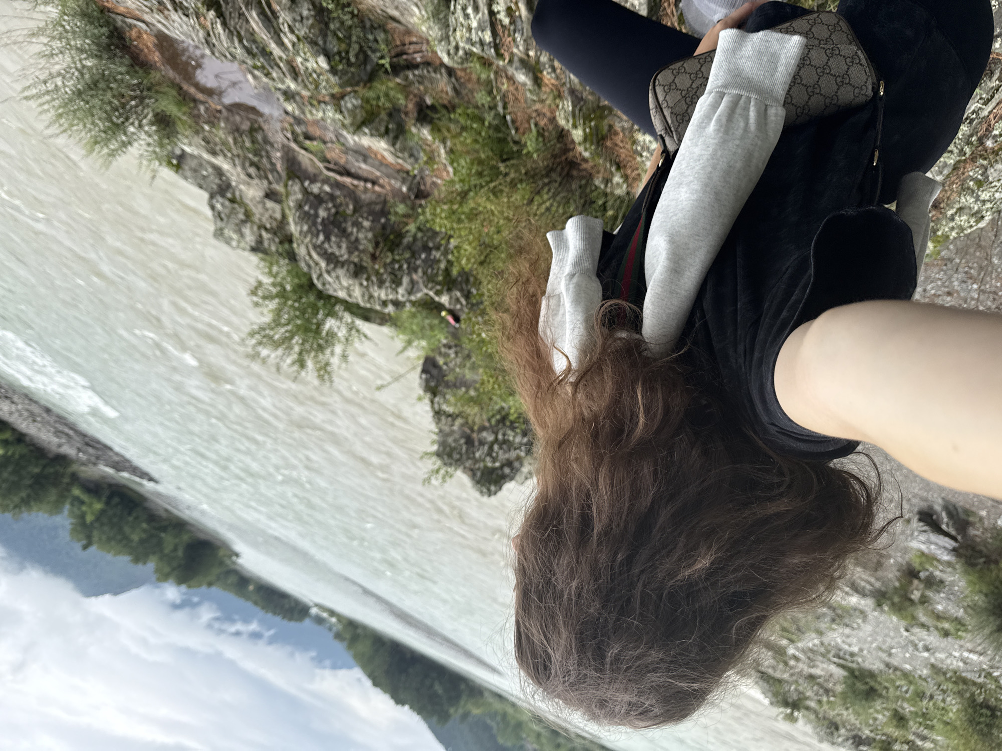 A person with long, dark, curly hair is standing on a rocky outcrop overlooking a wide, turbulent river. The person is wearing a black t-shirt with grey sleeves, dark leggings, and a small, patterned crossbody bag. The river water is a murky, light brown color and appears to be flowing quickly. The background shows a dense forest with green trees, and beyond that, mountains partially obscured by clouds. The weather appears overcast. The scene is located in Elekmonar, Russia.
