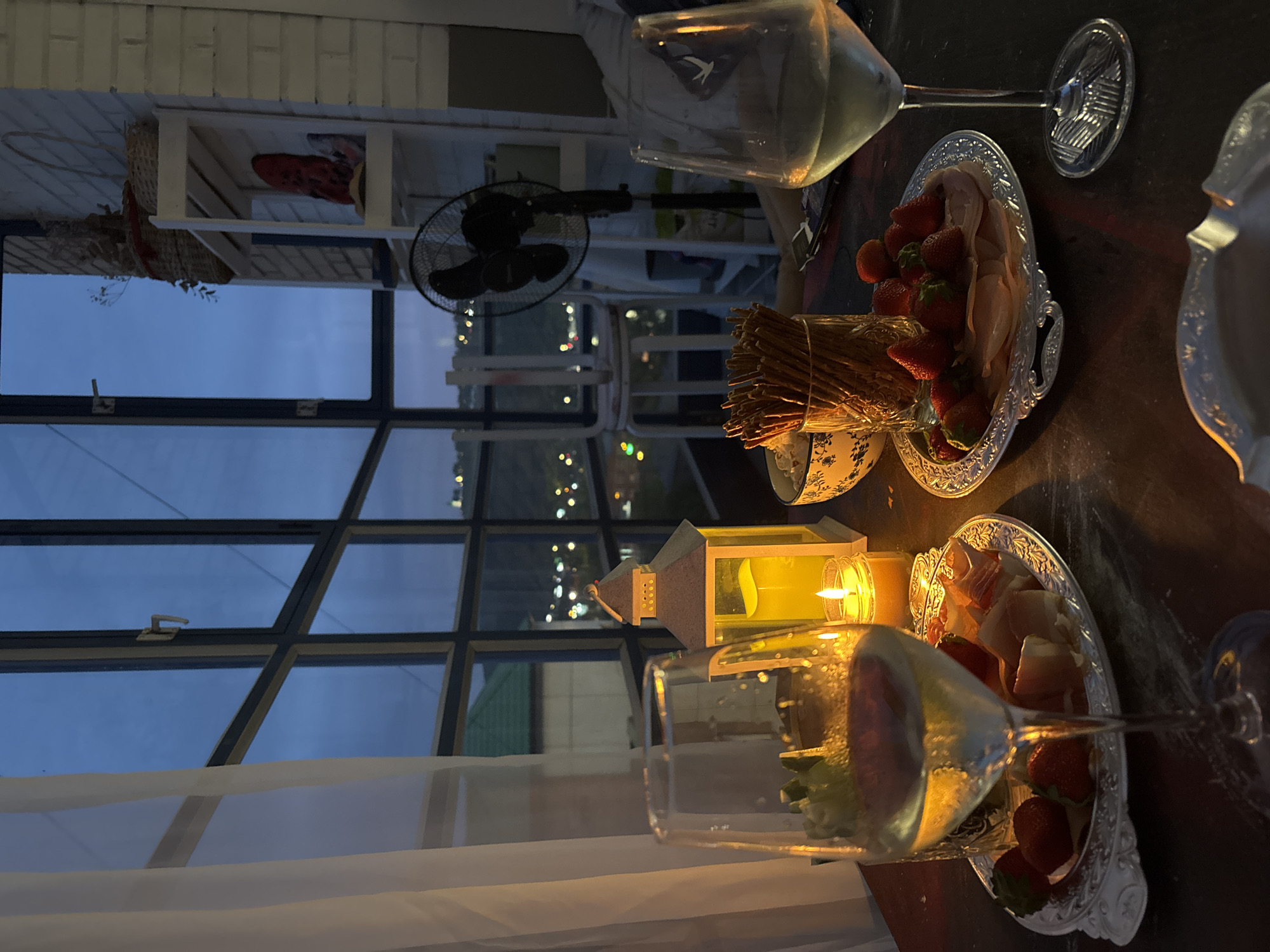Captured indoors, the image displays a dinner scene illuminated by the soft glow of candlelight, with trays of strawberries and prosciutto strategically arranged near the flickering flame. Wine glasses filled with golden liquid add a touch of elegance to the setting. A window reveals a cityscape, potentially identifying the location as Krasnoyarsk, Russia. A black electric fan stands in the background, close to a white shelf with a red object. The window is multi-paned with visible latch hardware, partially covered by a white curtain.