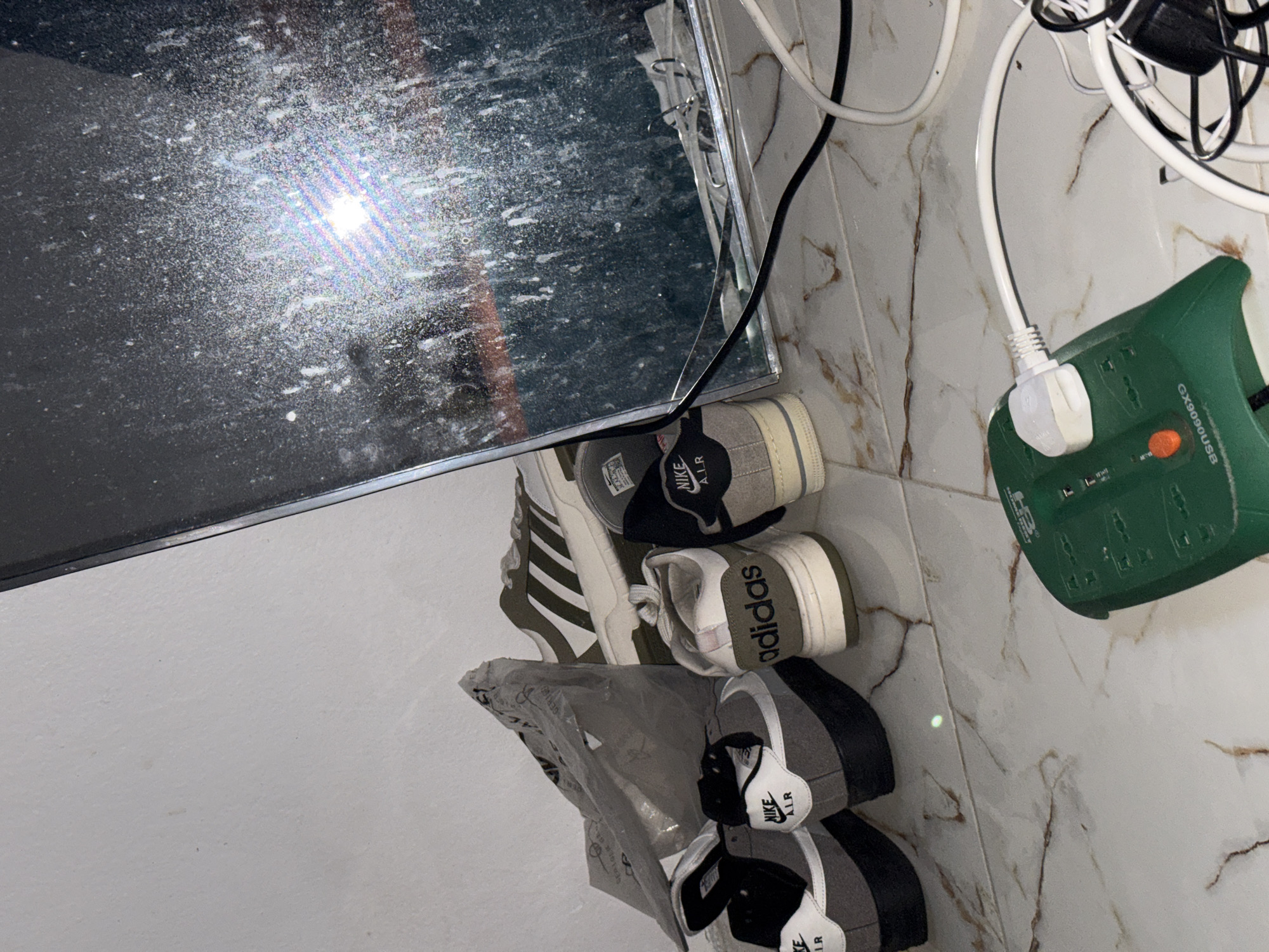 The image depicts a cluttered scene on a marble-patterned floor, likely indoors, with a white wall in the background. Several pairs of athletic shoes are visible, including Nike Air Force 1s and Adidas sneakers. A dirty, reflective surface, possibly a television screen, occupies the right side of the frame, displaying numerous smudges and a bright light reflection. Electrical equipment, including a green power strip with multiple outlets and various cords, is scattered on the floor. The overall impression is one of disarray and neglect, with no clear indication of specific human activity or interaction within the frame. The image does not provide any specific clues to pinpoint a location within Uyo, Nigeria, as the elements are generic and could be found in many urban environments.