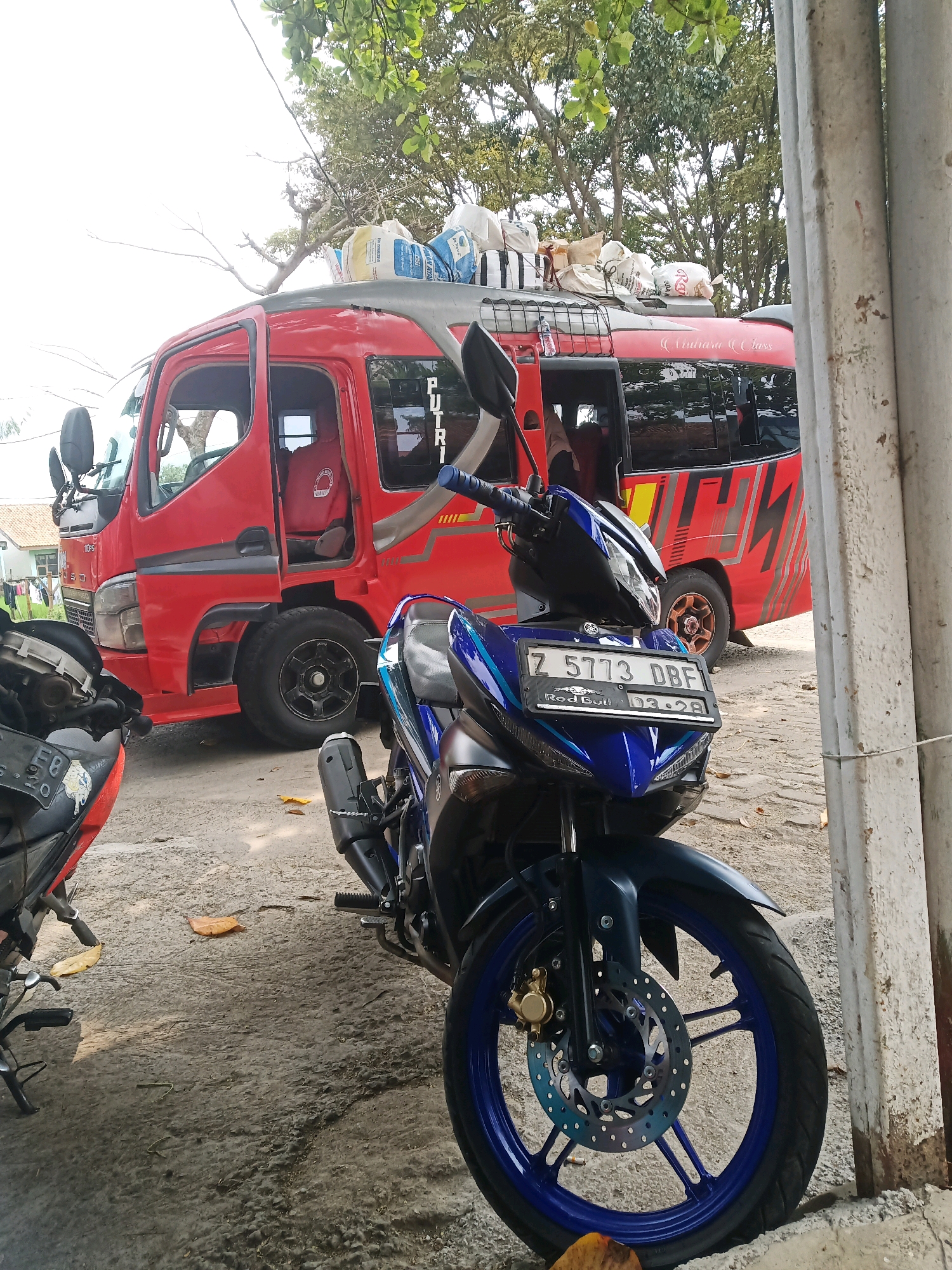 The image depicts a street scene in Garut, Indonesia, with two parked motorcycles in the foreground and a red bus in the background. The motorcycle on the right is blue and black, with a visible license plate reading "Z 5773 DBF". It has a "Red Bull" sticker and an expiration date of "03-28". The motorcycle on the left is partially visible and appears to be red and black with a license plate starting with "F8".
The red bus is parked behind the motorcycles. It has a cage-like structure on its roof carrying various bags and parcels. The side of the bus has a graphic design with red and yellow stripes and text that includes "PUTRI" and "Vitara Class". The bus appears to be a passenger vehicle, possibly a shuttle or tour bus.
The environment suggests an outdoor setting, likely a roadside or parking area, with a rough, paved ground surface. Trees with green foliage are visible in the background, and some fallen leaves are scattered on the ground. The lighting indicates it is daytime. The overall scene captures a moment of transit or waiting in a town or city in Indonesia.