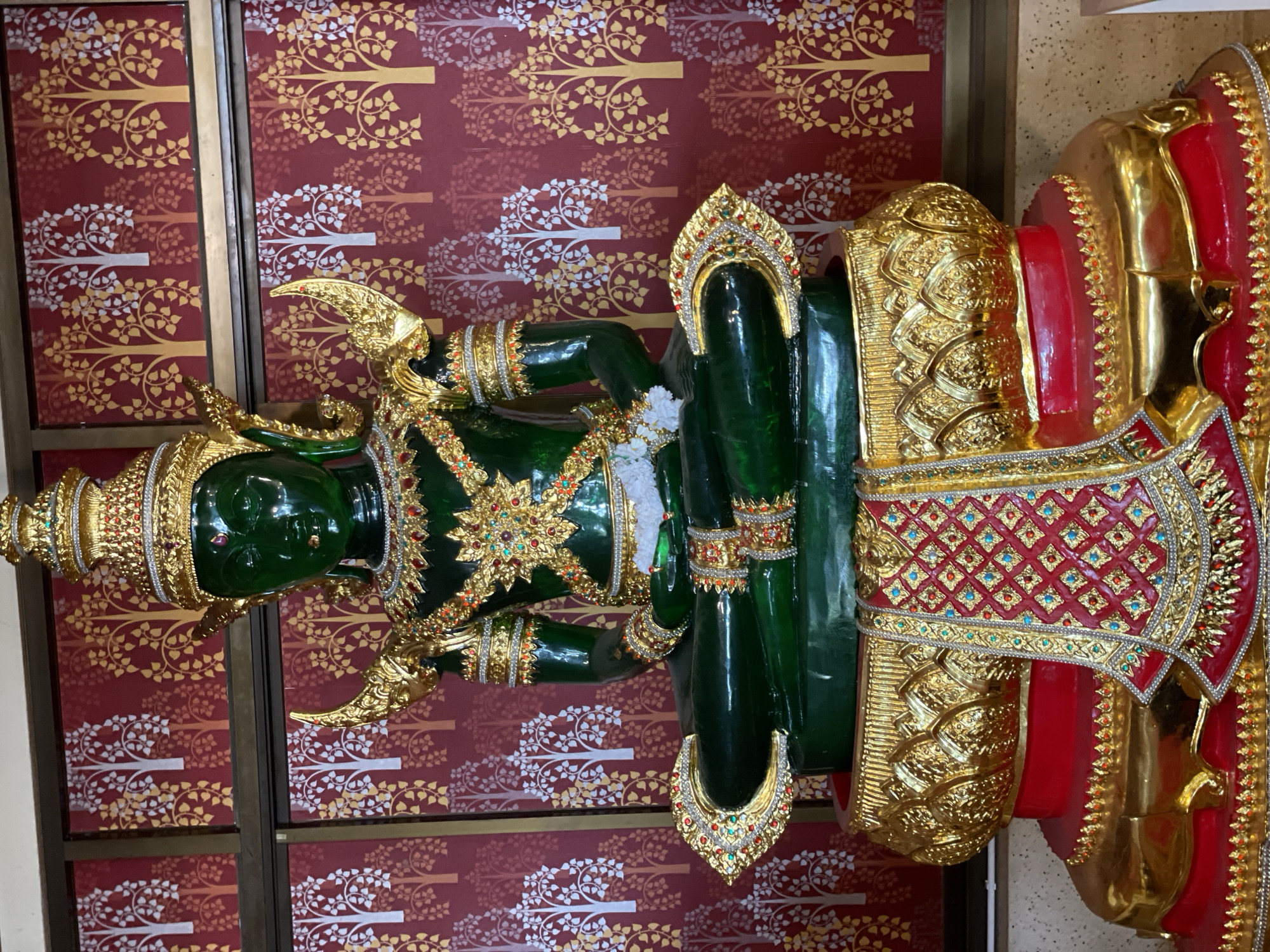 A full shot captures a seated, green-hued statue of a deity, likely Buddha, adorned in opulent gold and jewel-encrusted regalia, including a tall, tiered crown, ornate shoulder pieces, and intricate chest and arm embellishments. The statue is seated on a multi-tiered golden and red pedestal that features floral and diamond patterns. A richly decorated cloth, red with gold trim and a repeating diamond pattern of red and turquoise, drapes across the front of the pedestal. The background consists of red wallpaper with a repeating pattern of stylized trees in white and gold. The image appears to be taken indoors, possibly in a temple or shrine in Tambon Na Chom Thian, Thailand, given the stylistic elements and context of such imagery.