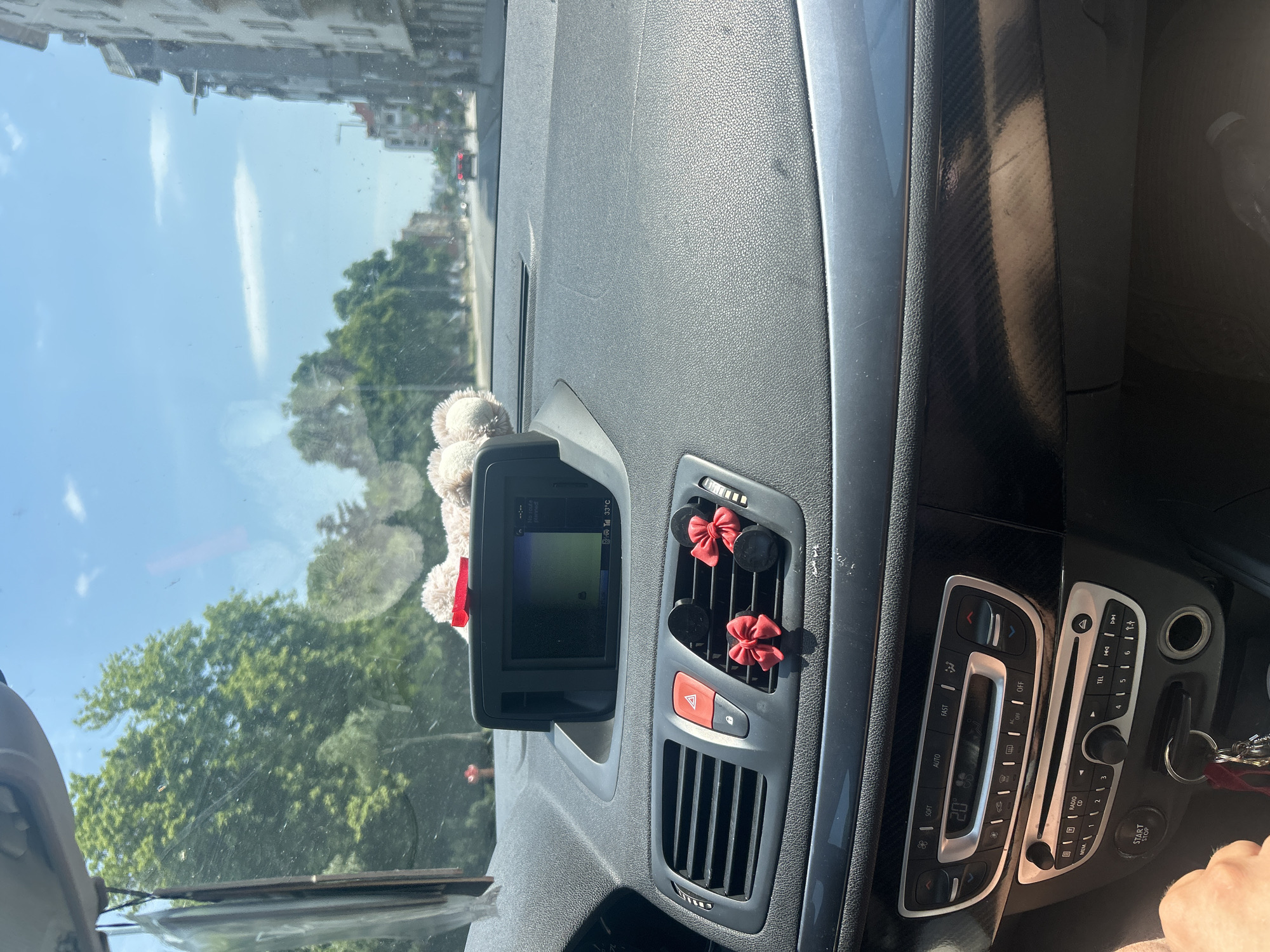 The image is a POV shot from inside a car, looking forward through the windshield. The car is driving on a city street lined with trees and buildings. The dashboard of the car is visible, with a navigation screen displaying a map, air vents, and the car's radio and climate controls. Two small red bows are attached to the air vents, and a stuffed animal toy is visible on top of the dashboard. The sky is blue with a few white clouds, indicating it is daytime. The scene suggests the car is moving through a moderately busy urban area, possibly Plovdiv, Bulgaria, given the presence of architectural styles common in Eastern Europe.