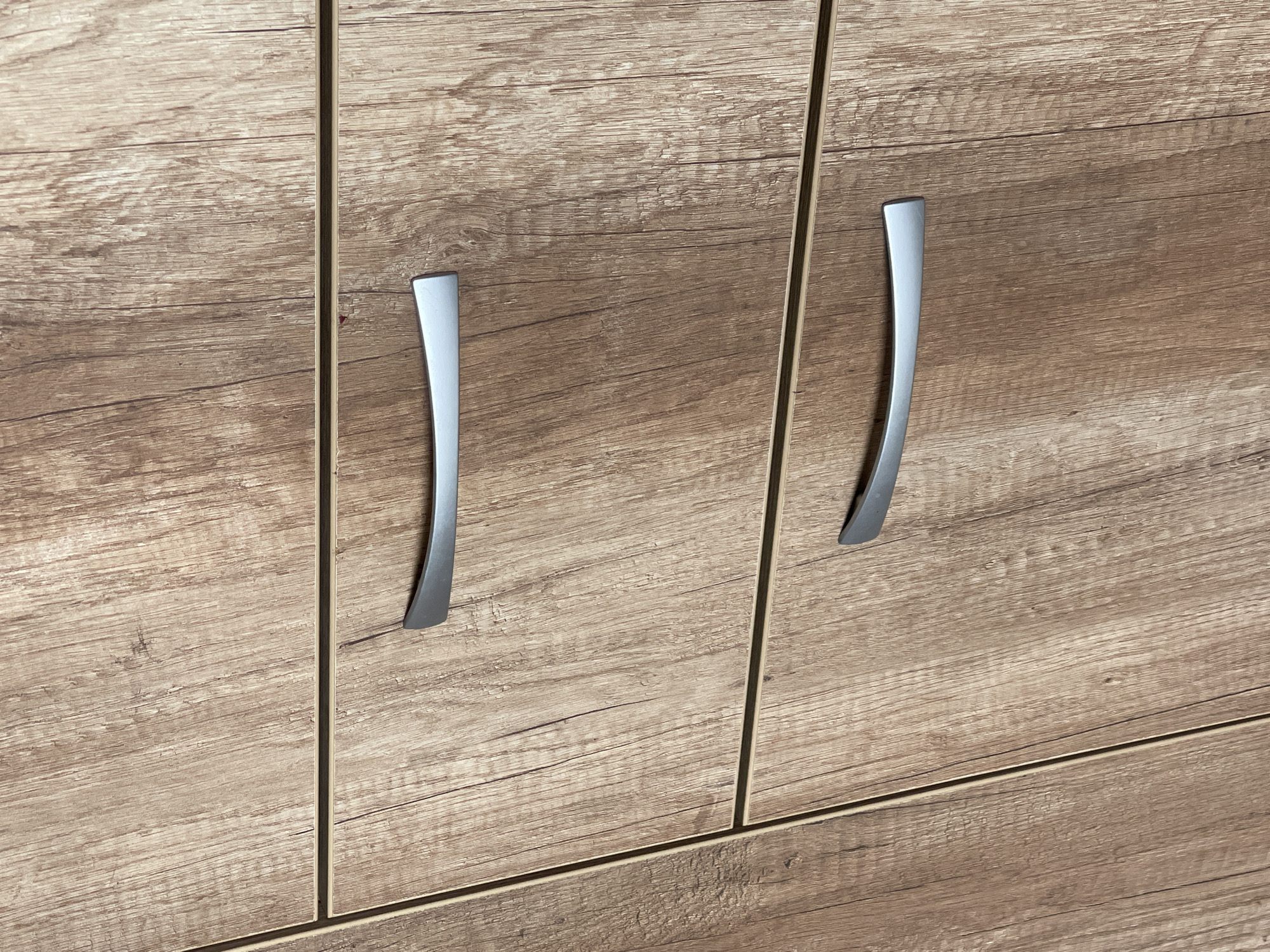 The image shows a close-up of a set of wooden cabinet doors with curved, silver-colored handles. The wood has a light brown, textured finish, resembling wood grain. The handles are vertically oriented, positioned towards the edges of the doors. The setting is indoors. Somovit, Bulgaria is not relevant to the image.