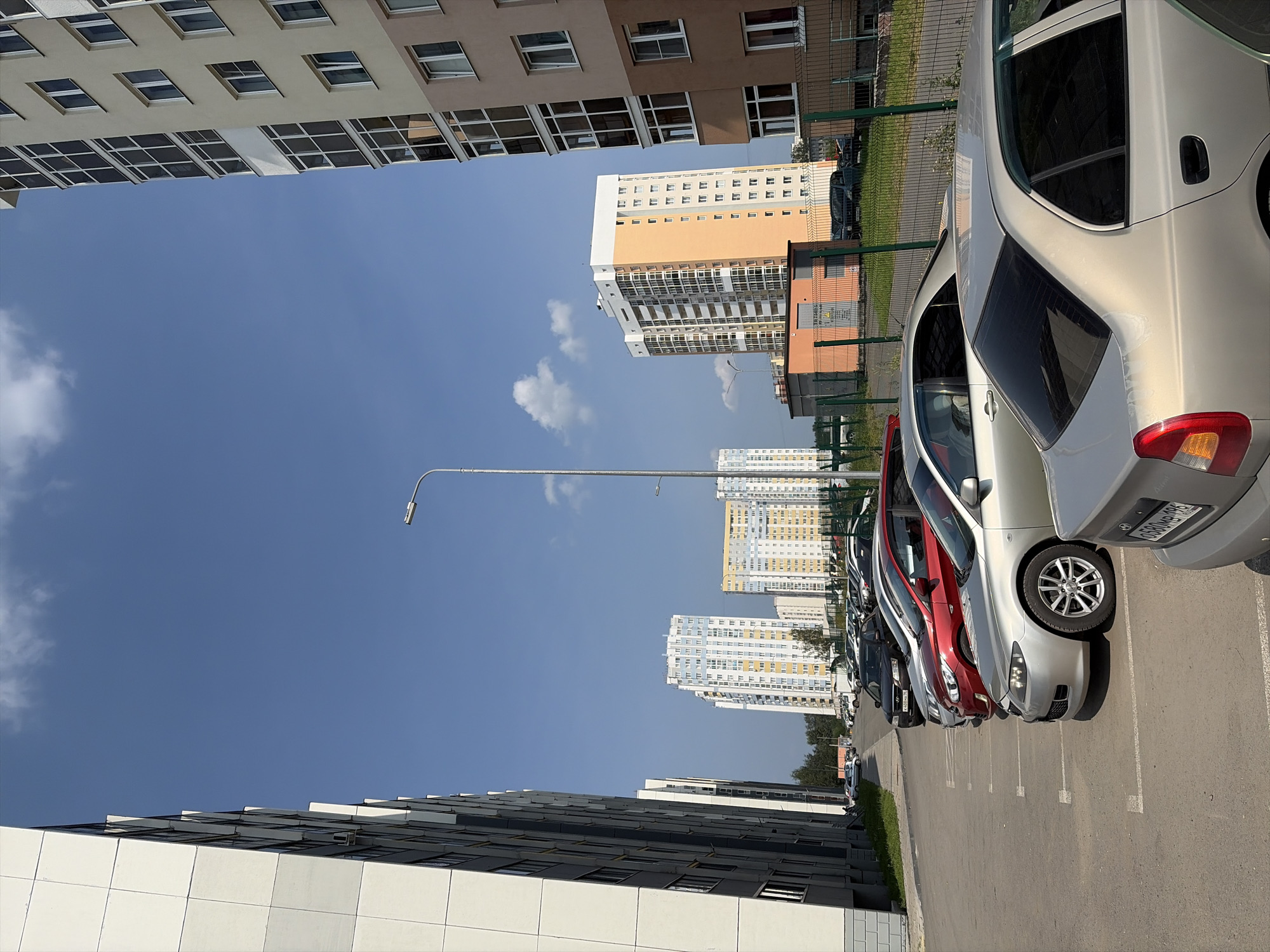 The image depicts a parking lot in a residential area of Yekaterinburg, Russia. Several multi-story apartment buildings dominate the skyline, with a mix of light-colored facades and some with orange accents. A row of parked cars, predominantly sedans and hatchbacks, fills the paved lot. Among the visible vehicles are a silver sedan in the foreground, a red hatchback behind it, and a black car further down the line. A lamppost stands to the right of the frame, and a green metal fence encloses a grassy area beyond the parking lot. The sky is a clear blue with a few scattered white clouds, suggesting fair weather. The overall scene conveys a typical urban residential environment.