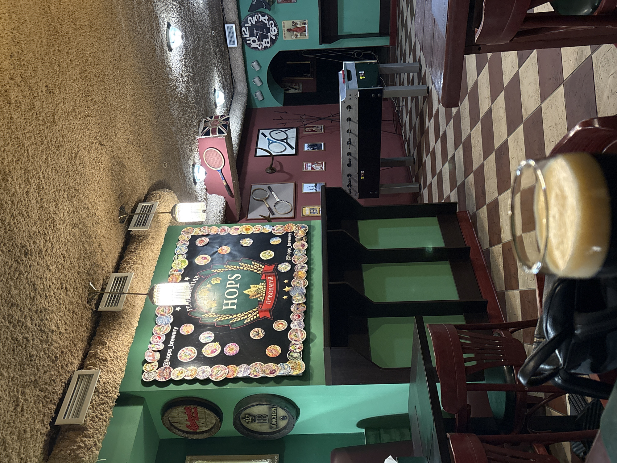 The image depicts the interior of "Hops Brewery" in Minsk, Belarus. The walls and ceiling are textured with a beige, somewhat shaggy material. A large, illuminated sign advertising the brewery features the word "HOPS" in Cyrillic and English, surrounded by various beer-related imagery. Below the sign are decorative beer barrels with brand names. A foosball table is positioned near the back, close to a wall adorned with tennis rackets and sporting memorabilia. A brown and white checkered floor leads towards the back of the establishment. In the foreground, a glass of beer with a visible head is placed on a table, with a dark leather jacket draped over a red wooden chair. The room has green walls and is lit with a combination of spotlights and decorative fixtures. The overall ambiance suggests a brewery-themed pub or restaurant.