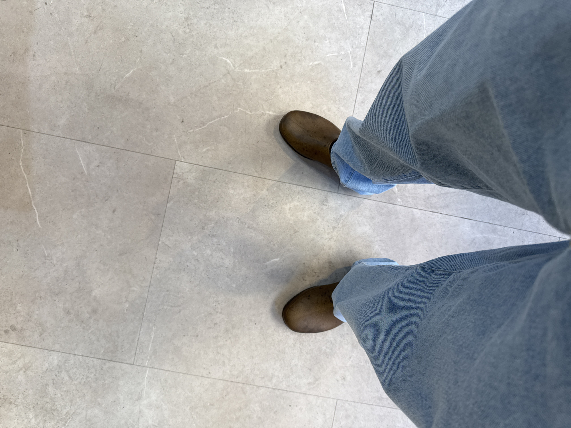 An overhead, close-up shot captures a person's lower legs and feet, seen from a first-person perspective. The individual is wearing light-wash blue jeans that are slightly cuffed or bunched at the ankles. They have on dark brown, low-cut shoes or boots, possibly made of leather or a similar material.
The person is standing on a light grey tiled floor, composed of large rectangular tiles. These tiles feature subtle, irregular white veining, suggesting a natural stone or marble effect. Thin, dark grout lines delineate the individual tiles, creating a grid pattern across the surface. The lighting is even, indicative of an indoor or covered outdoor environment, a common sight in urban settings like London, United Kingdom. No other subjects, objects, or text are visible in the frame, and the scene appears static, depicting a moment of standing.