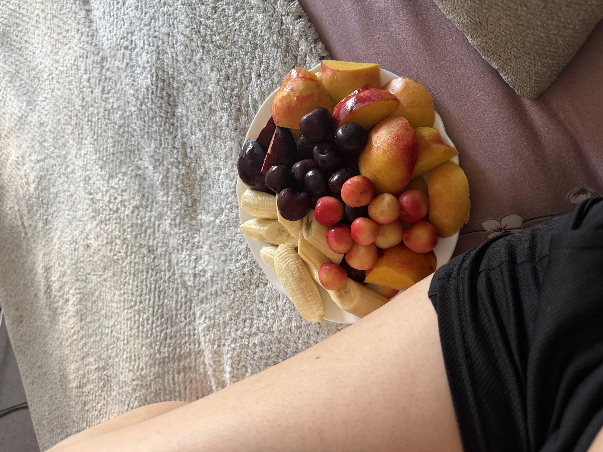 A white plate filled with sliced fruits including bananas, cherries, peaches, and plums is positioned on a textured, light-colored surface. Partially visible in the lower left of the image is a person's bare leg and thigh, clad in black shorts. The background consists of a soft, neutral-colored textile. There is no information in the image to suggest the location is Tbilisi, Georgia.