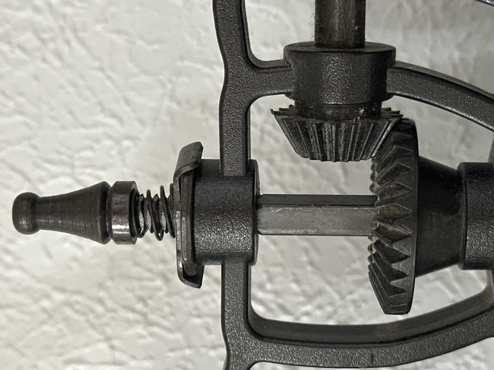 The image shows a close-up, detailed view of a metal mechanism, likely part of a vintage hand-cranked device such as a coffee grinder or a meat grinder. The focus is on the gears and internal workings. A central vertical shaft is visible, topped with a cap and a coiled spring. Below this, a large bevel gear with serrated teeth is mounted. Intersecting this gear is a smaller bevel gear, also with teeth, attached to a horizontal shaft. The metal components are dark and appear to be cast iron or a similar material, exhibiting a textured surface. The background is a textured, light-colored wall, possibly stucco or wallpaper, which is out of focus. There are no people, animals, or any indications of a specific city or location, including Nevinnomyssk, Russia. The lighting is even, highlighting the metallic textures and intricate details of the mechanical parts.