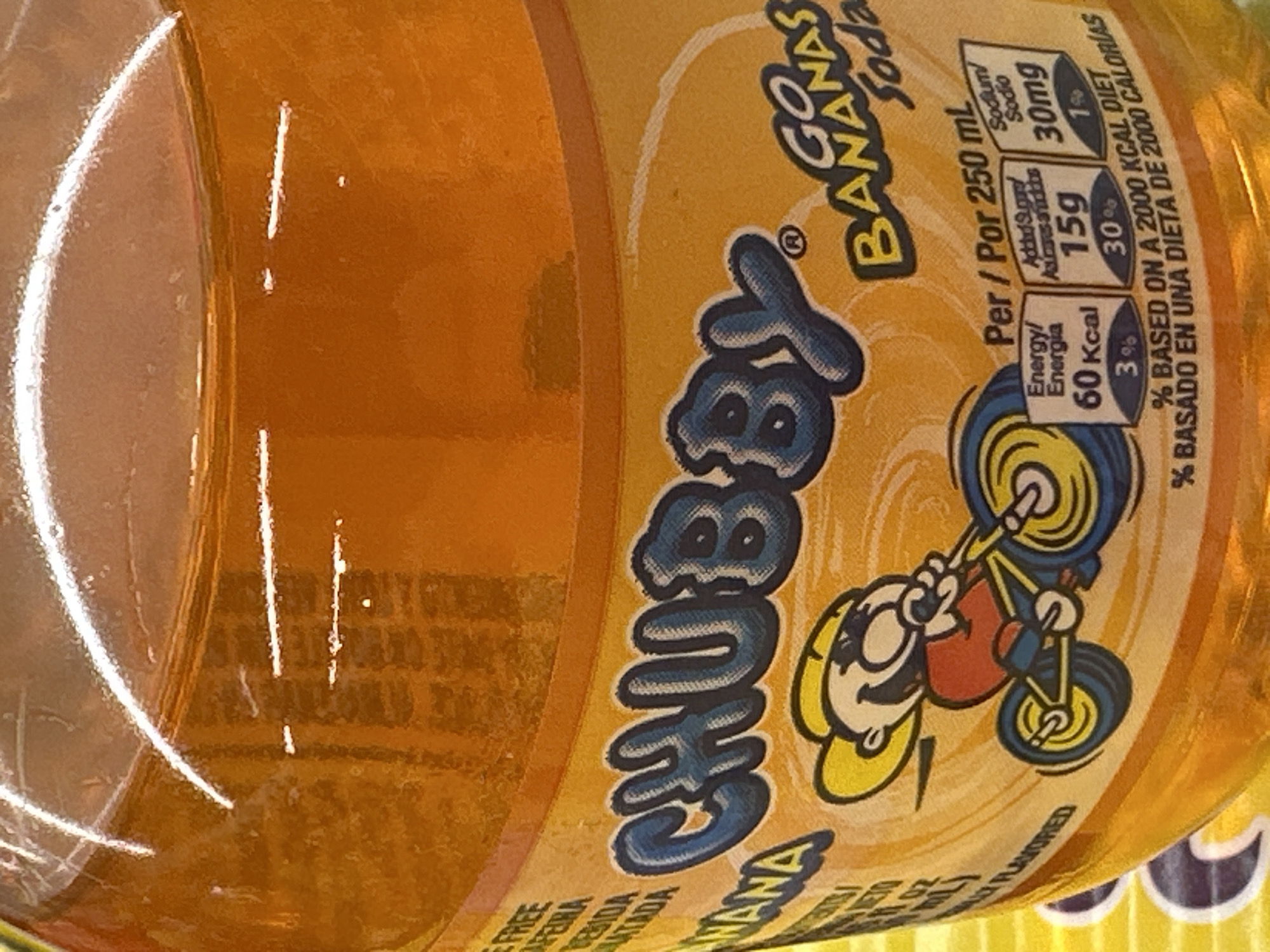 The image shows a close-up of a bottle of "CHUBBY Go BANANAS Soda". The bottle is filled with a golden-colored liquid. The label features a cartoon character riding a bicycle and nutritional information. The label also includes text in English and Spanish. The background is a blurry orange and yellow color. There is no visible indication of the city of Camp Hill, United States of America in this image.