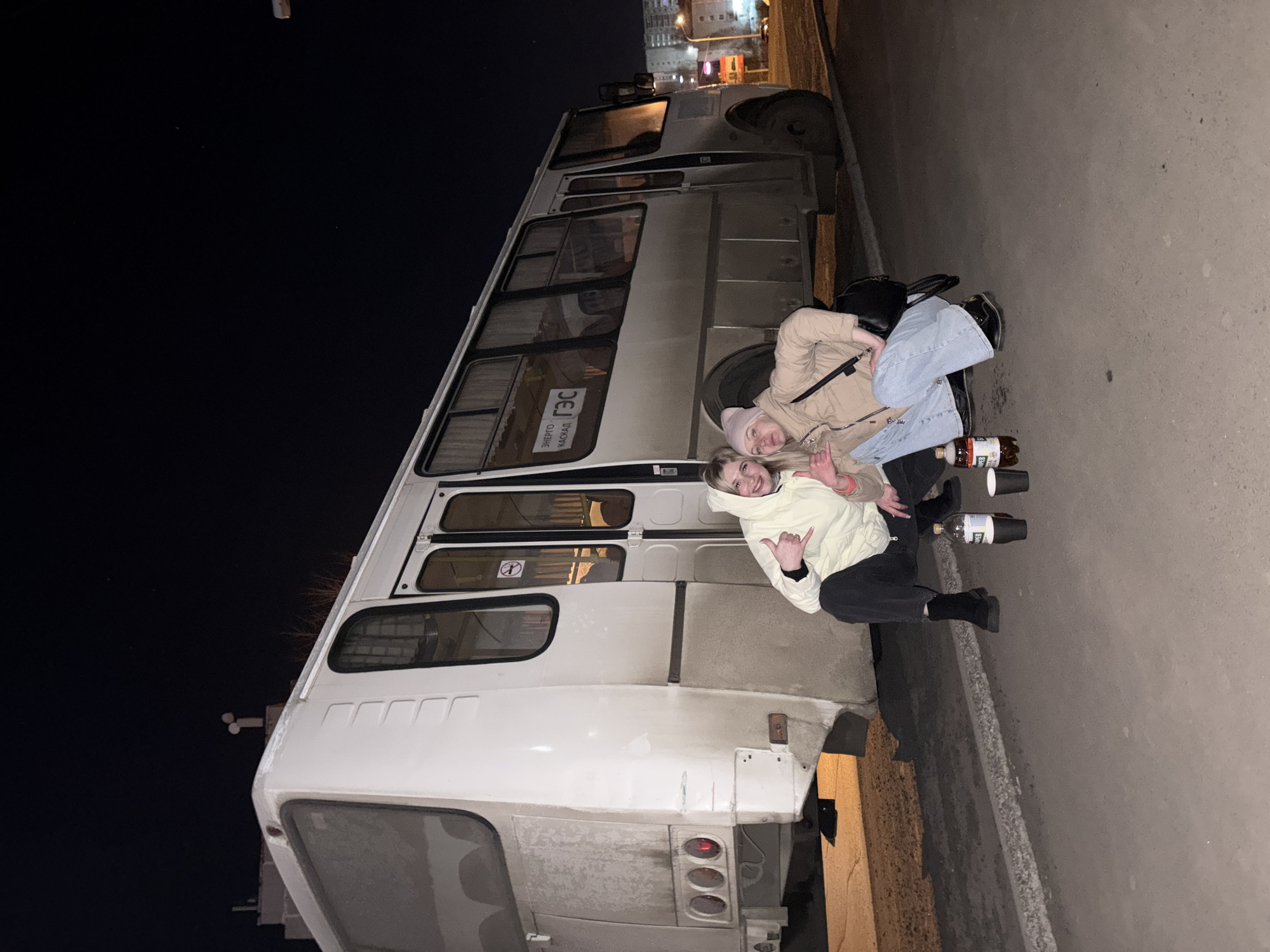The image depicts two young women sitting casually on the ground next to a white minibus or bus at night. The photograph is oriented vertically, rotated 90 degrees clockwise from a typical horizontal shot.
The scene takes place outdoors on what appears to be a paved surface, possibly a street or sidewalk, with a large, white bus parked alongside. The background is dark, indicating nighttime, with distant artificial lights from buildings visible.
One woman, on the left, is wearing a light brown or tan jacket, light blue wide-leg jeans, and a pink beanie. She has a black shoulder bag and is smiling directly at the camera. The second woman, on the right, wears a light yellow or cream-colored hooded sweatshirt and dark pants. She is also smiling and making a "shaka" (hang loose) hand gesture towards the camera. Both women appear relaxed and cheerful.
On the ground next to them are several items, including a large plastic bottle containing a dark liquid (possibly kvass or another beverage) and two disposable cups, one white and one darker, suggesting they are drinking together.
The bus is a passenger type, possibly a Russian Gazelle or similar model, featuring multiple windows along its side. On one of the bus windows, there's a visible "No Smoking" symbol. Cyrillic text is clearly visible on the side of the bus: "ЭНЕРГО" (ENERGO), "КАСКАД" (KASKAD), and "ГЭС" (GES), which likely refer to an energy company or hydroelectric power station, given the "ГЭС" abbreviation.
The overall mood is casual and social, captured in an urban or semi-urban setting in Ust-Ilimsk, Russia, under artificial light during the night.