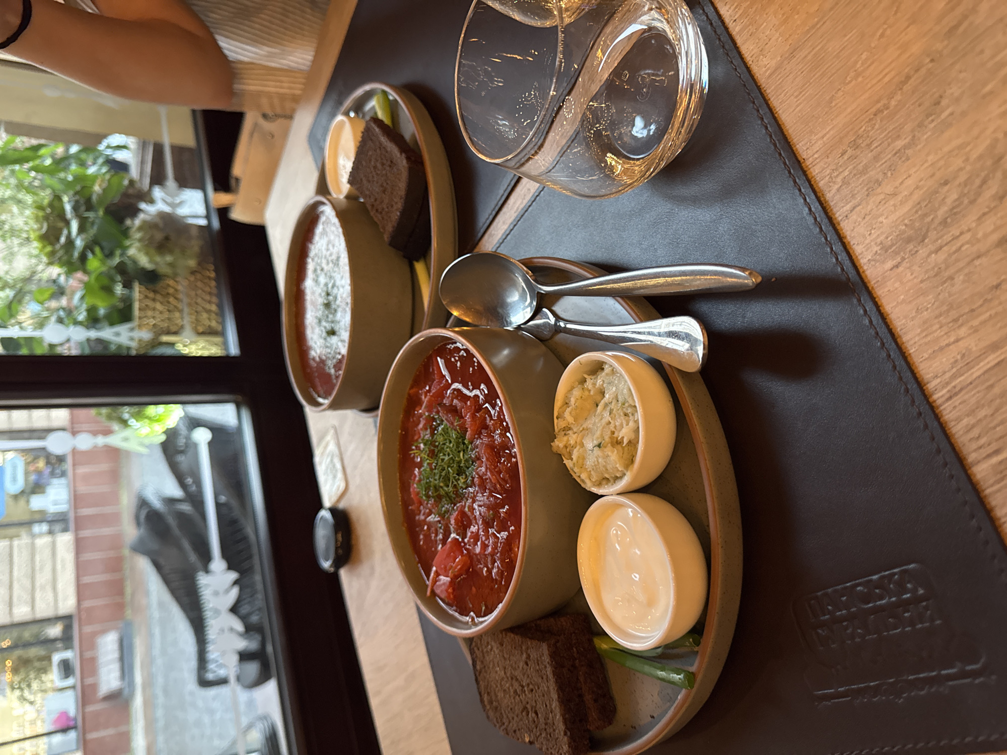 A close-up, eye-level shot shows two bowls of borscht on a table in Chernivtsi, Ukraine, likely at a restaurant given the setting. The borscht in the foreground is in a light green ceramic bowl, garnished with chopped green herbs. Beside it are two small white bowls, one with a dollop of sour cream and the other with a spread, likely horseradish or a similar condiment. Two slices of dark rye bread and a stalk of green onion are also on the plate. A spoon rests on a dark brown placemat. In the background, another bowl of borscht is visible on a separate plate, with a slice of bread and a small portion of sour cream. Beyond the food, a large window offers a view of a street scene with a glimpse of buildings and greenery. The table appears to be made of wood. A clear glass, possibly containing water or a beverage, is positioned to the right of the foreground plate. The overall lighting is warm and inviting, suggesting an indoor dining setting.