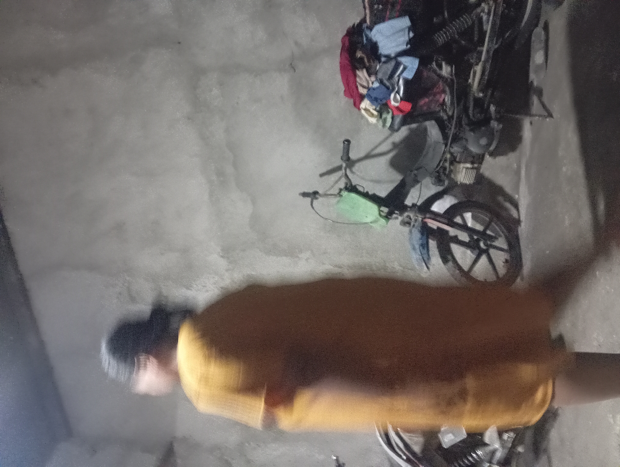 In Rejosari, Indonesia, a person, blurred by motion, stands in what appears to be an indoor or covered outdoor space with a rough, concrete wall as a backdrop. The individual is wearing a long, loose-fitting yellow garment. To the right of the person, a motorcycle is parked, its rear section piled high with various articles of clothing, including red, blue, and light-colored fabrics. Another motorcycle is partially visible to the left of the person. The lighting is dim, creating shadows and a slightly grainy texture throughout the image.