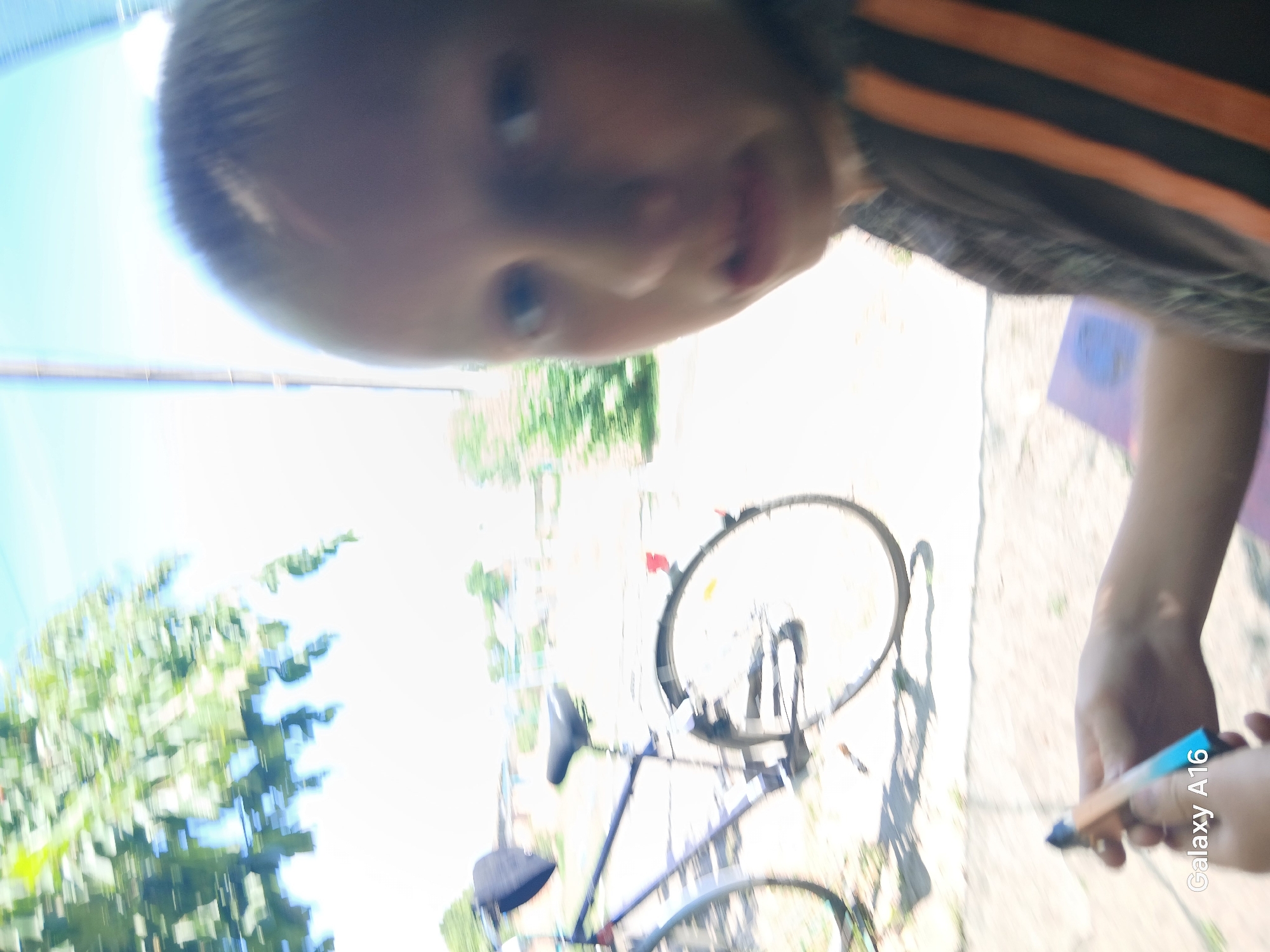 The image depicts a blurred outdoor scene, likely in a rural or suburban area, with a child in the foreground and a bicycle in the mid-ground. The child, partially obscured by motion blur, appears to be holding a small, colorful object, possibly a pen or a vaping device. The child is wearing a dark shirt with orange stripes. Behind the child, a bicycle with a black seat and fender is visible. The background consists of trees, buildings, and a bright sky. The "Galaxy A16" watermark indicates the image was taken with a Samsung Galaxy A16 smartphone. The specific location of Ustyvytsia, Ukraine, is not visually discernible from the image itself.