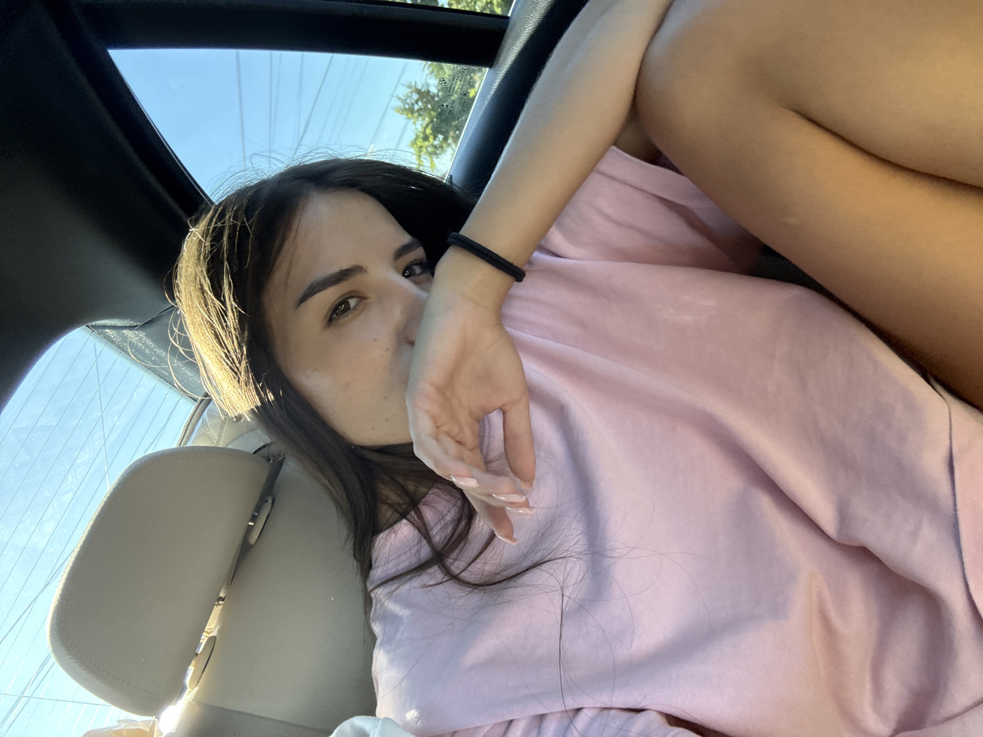 A young woman with long brown hair and fair skin is seated in the back of a vehicle, her right arm resting across her chest and her hand covering her mouth. She is wearing a pink t-shirt and appears to be looking directly at the camera. The background shows the interior of a car, including headrests and windows. Through the windows, a bright blue sky and trees are visible, suggesting an outdoor setting. There are no discernible landmarks that would place the image specifically in the city of Zorleni, Romania.