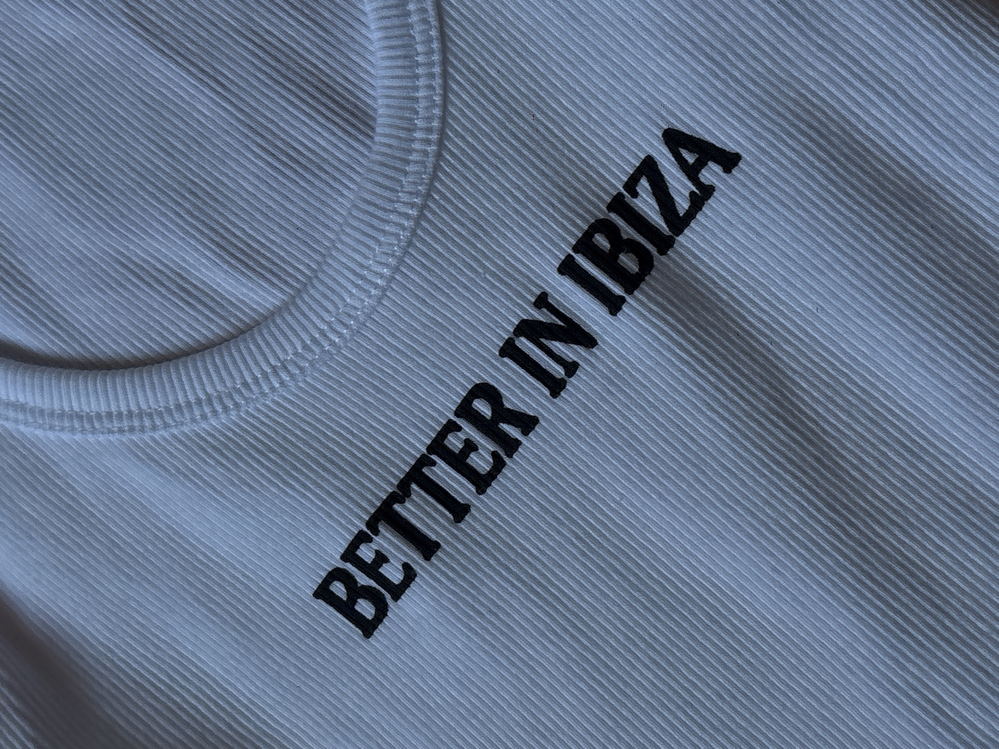 The image shows a close-up of a white ribbed tank top with the text "BETTER IN IBIZA" embroidered in black. The top is lying flat, and the neckline is visible in the upper left. The ribbed texture of the fabric is evident. No people, actions, or environmental details are visible. There is no information to suggest the photo was taken in Mexico City, Mexico.