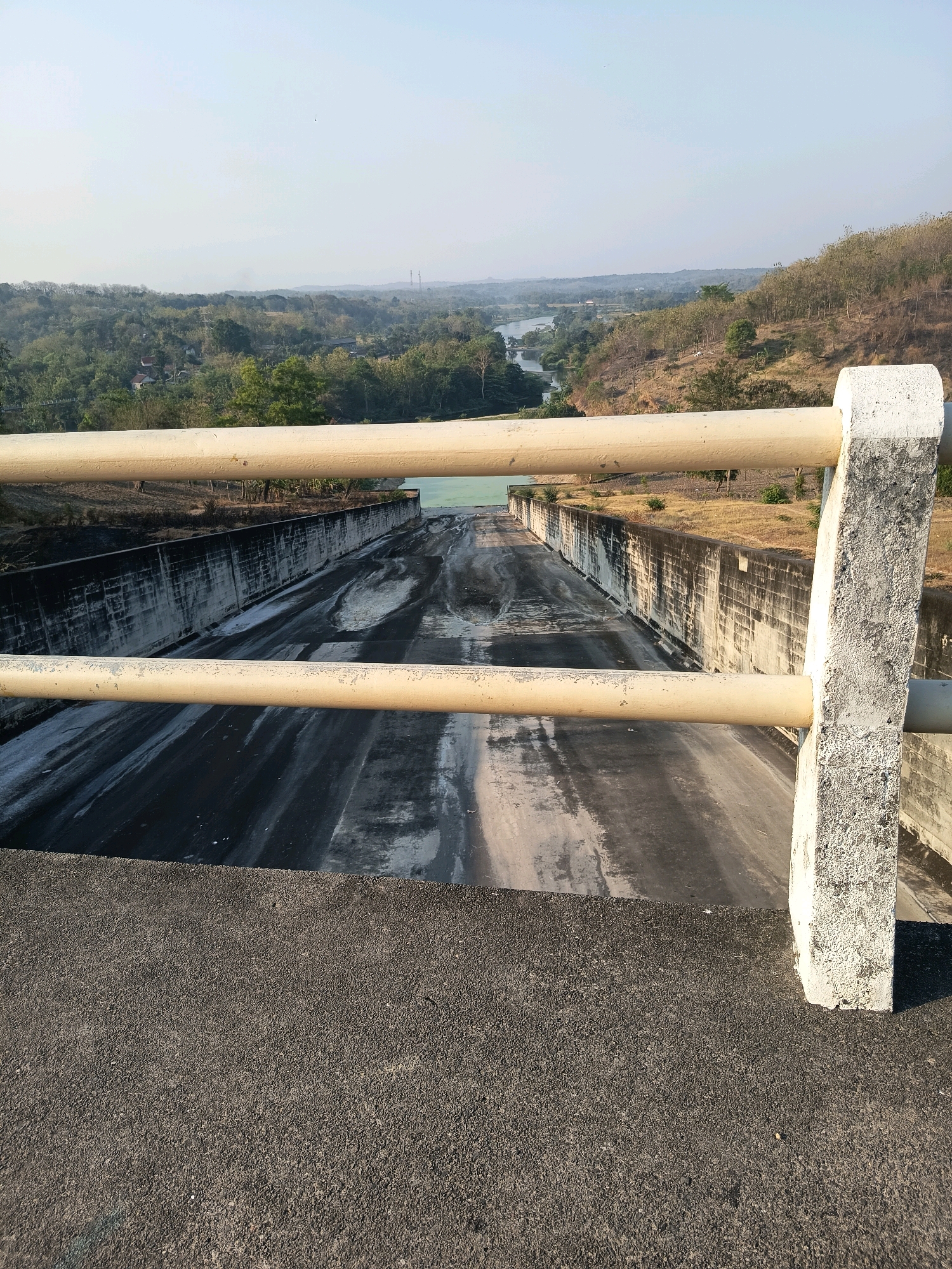 The image shows a view from a dam spillway, looking towards a river and a hilly landscape. The spillway itself is a long, sloping channel with concrete walls on either side. The surface of the spillway is dark gray with lighter streaks and patches, suggesting water flow or residue. Two horizontal, light-colored railings are visible in the foreground, with a concrete post on the right. Beyond the spillway, a calm river winds through a valley. The banks of the river and the surrounding hills are covered with lush green vegetation, with some dry patches on the steeper slopes. The sky is clear and pale blue, with a hazy horizon. The overall impression is of a man-made structure integrated into a natural landscape. The location is identified as the Kalangbancar Dam in Kalangbancar, Indonesia. There are no people or animals visible in the image.