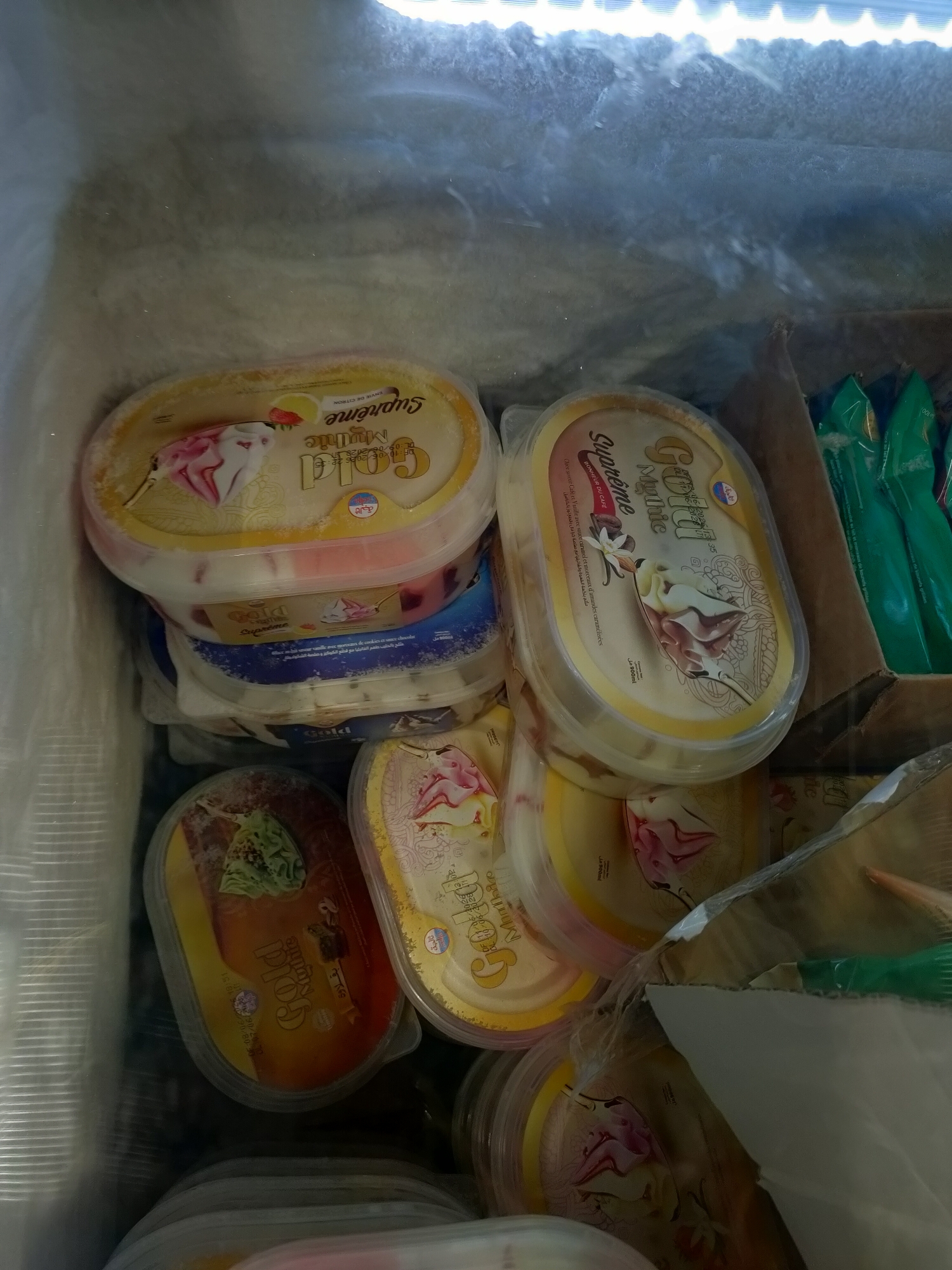 In the city of Unknown, Tunisia, a freezer is filled with multiple tubs of Gold Supreme ice cream. The tubs are oval-shaped with yellow lids. Some lids display images of ice cream and floral designs, with text in what appears to be Arabic and French, including "Gold Supreme," "Mythic," and "Bonheur Du Cap." The ice cream tubs are stacked on top of each other, with some partially obscured by plastic wrap and other packaging. In the background, green packaging for what appears to be other frozen products is visible, along with a brown cardboard box. The freezer is frosted over, indicating a cold environment.