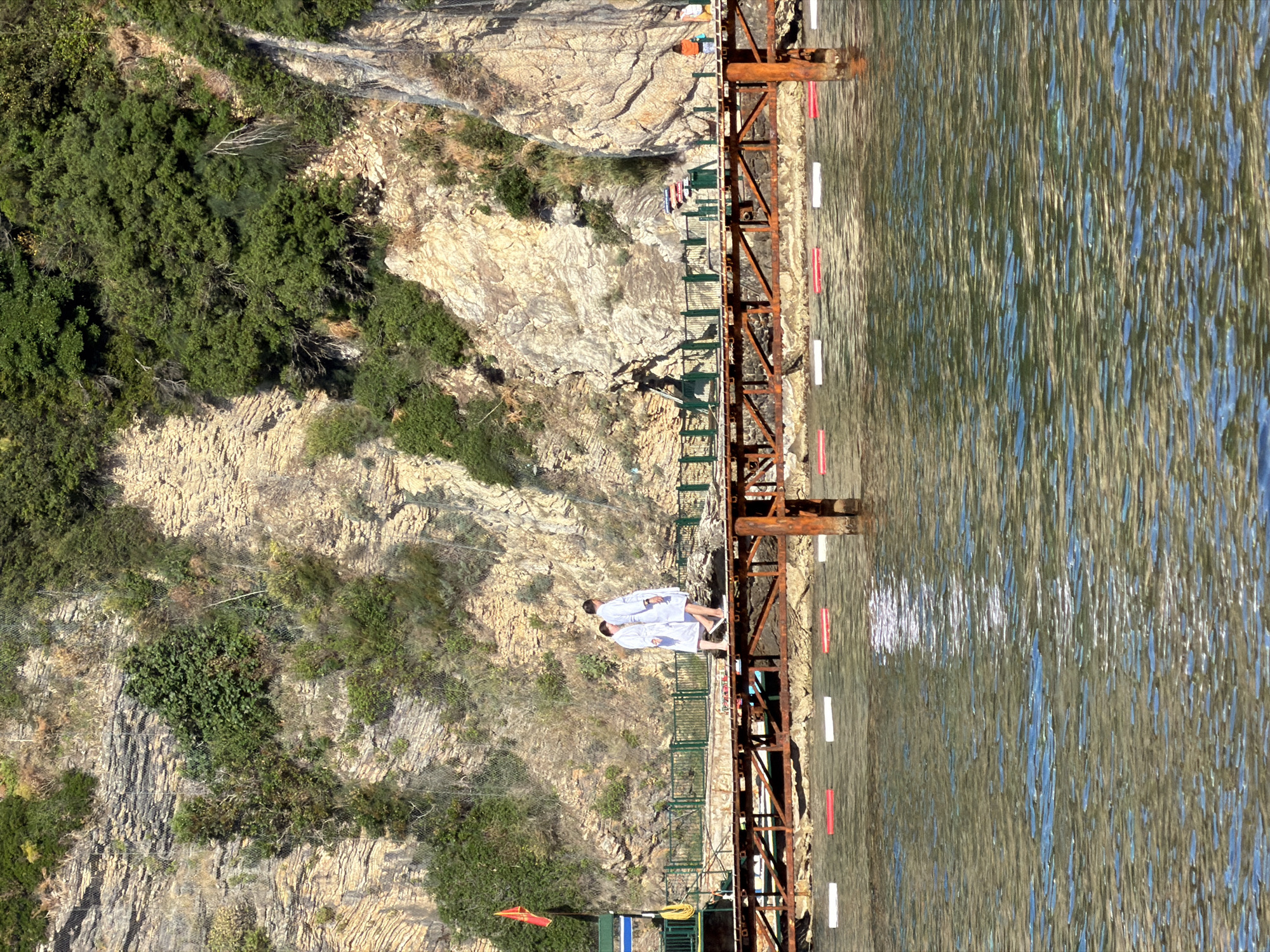 The image depicts a coastal scene in Budva, Montenegro, featuring a rocky cliff face covered with vegetation and a section of the Adriatic Sea. A rusty metal structure with a green railing extends along the edge of the cliff into the water. Two individuals wearing white robes and footwear stand on the walkway of this structure. The sea displays a textured surface with reflections of the sky.