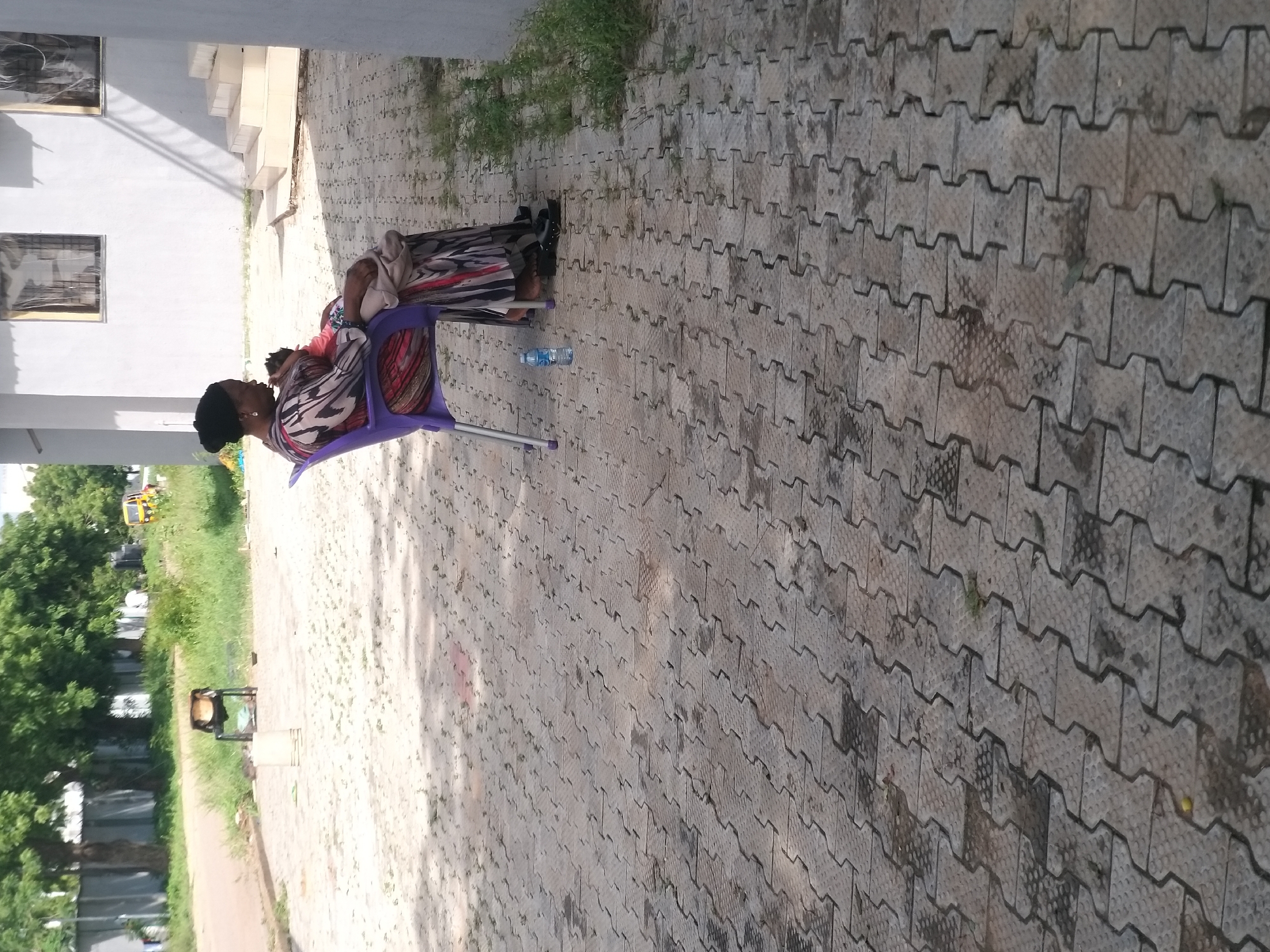 A woman wearing a black headscarf and a colorful patterned dress is sitting on a purple plastic chair in front of a white building. She is holding a baby in her arms, with the baby's face obscured by her arm. A bottle of water is on the ground next to the chair. The ground is paved with interlocking grey bricks. In the background, there are trees and a small yellow vehicle. This scene is likely located in Kano, Nigeria, given the common architectural style and climate.