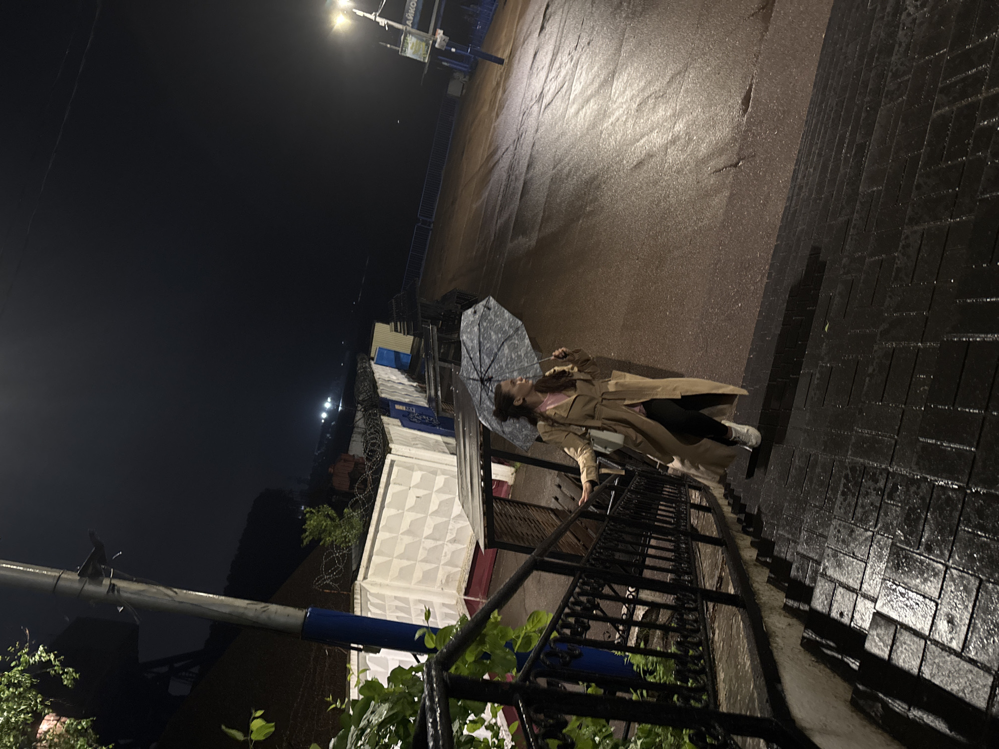 The image depicts a nighttime outdoor scene in the city of Chaikovsky, Russia, characterized by wet surfaces indicative of recent rain. A young woman, dressed in a light-colored trench coat, pink top, dark trousers, and white sneakers, is descending a set of wide, dark, wet paved stairs. She holds an open, patterned umbrella in her left hand, angled away from her body. Her right hand is near a black ornate metal railing that borders the stairs on the left.
To the left of the stairs, green foliage is visible, and a utility pole with a blue base stands upright. In the mid-ground, behind the railing and foliage, there is a white concrete wall with a geometric diamond pattern, topped with barbed wire fencing. Beyond the stairs to the right, a wide, upward-sloping asphalt or paved road appears wet and reflective. A prominent street light on the right side of the road illuminates the area, casting reflections on the wet ground. Near this streetlight, a sign is visible with partial Russian text "ЧАЙКО", consistent with "ЧАЙКОВСКИЙ" (Chaikovsky). The distant background shows faint lights, suggesting further urban infrastructure. The overall environment is dark, illuminated primarily by artificial street lighting.