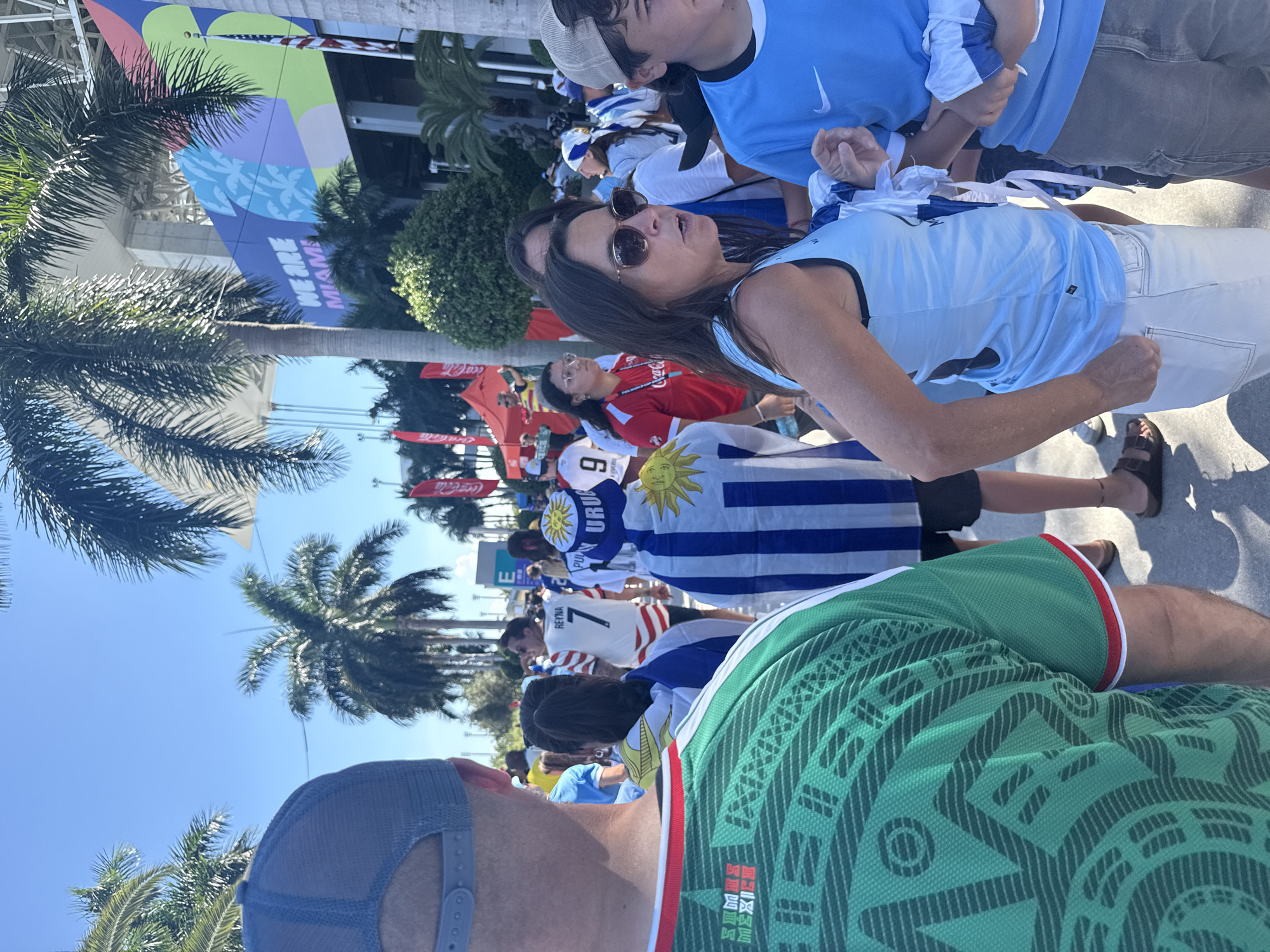 This image depicts a crowd of people gathered outdoors, likely for an event. The subjects are varied, with individuals wearing jerseys and hats, suggesting a sporting event or festival. One prominent detail is the banner in the background with text that reads "WE ARE MIAMI," indicating the location as Miami. The clear blue sky and palm trees further support a warm, sunny environment, suggesting daytime.
Several individuals are wearing what appear to be soccer jerseys with national flags or symbols. One person is wearing a light blue jersey with a Nike logo, and another is seen in a green jersey with a pattern that resembles traditional Mexican designs. Flags, including one with blue and white stripes and a sun emblem, are visible, hinting at international participation.
The overall mood appears to be one of excitement and anticipation, with people looking engaged and some appearing to be in conversation. The presence of Coca-Cola signage in the background is a common element at large public events. The image captures a vibrant atmosphere, with a focus on the fans and their attire, showcasing their support for their respective teams or countries.