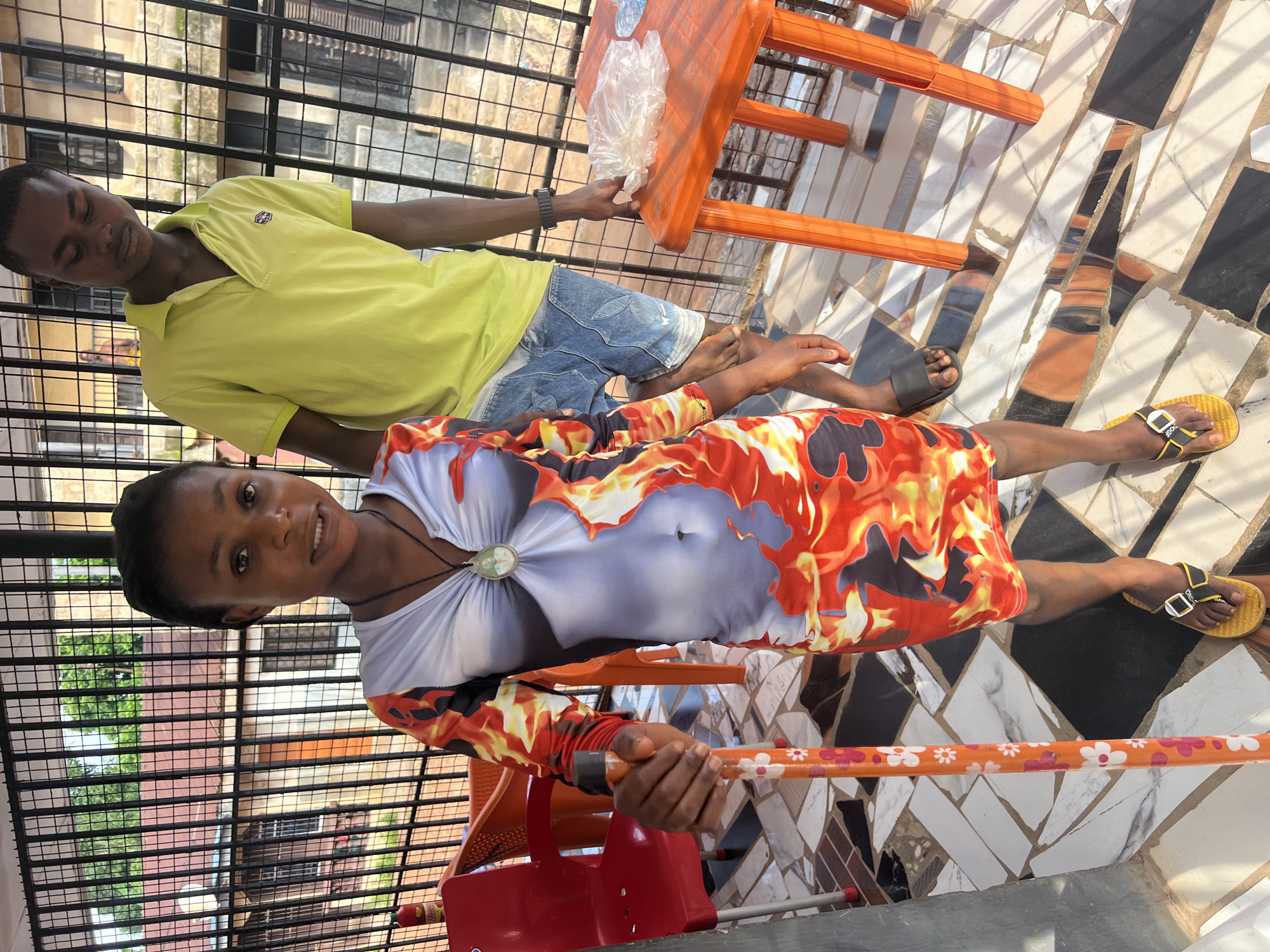 In Nkpor, Nigeria, a young girl stands on a tiled patio, holding an orange-colored walking stick decorated with white and pink flowers. She is wearing a long-sleeved dress with a fiery pattern of red, orange, and black. A dark necklace with a circular pendant hangs around her neck. To her right, a young man stands, wearing a lime green polo shirt and denim shorts. He appears to be looking down. In the background, a black metal fence encloses the patio, with buildings and trees visible beyond. An orange plastic table stands to the right of the man, with a plastic bag on top. Two red plastic chairs are visible on the left side of the image. The scene appears to be outdoors, possibly on a balcony or veranda.