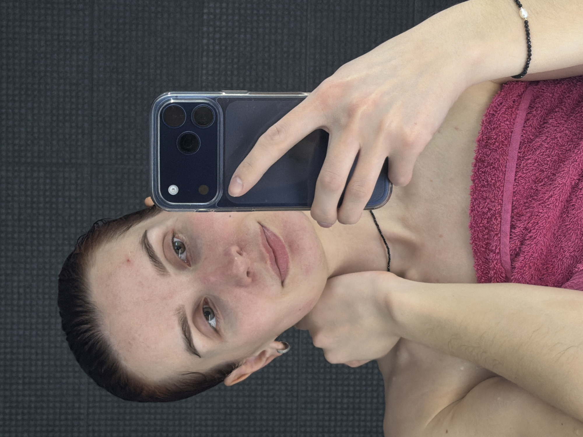 A person is taking a selfie with a smartphone in a room. The person has a towel wrapped around their body and is wearing a bracelet and a necklace. The background is a gray surface. The location is unspecified, and the reference to Chaikovsky, Russia, is irrelevant.
