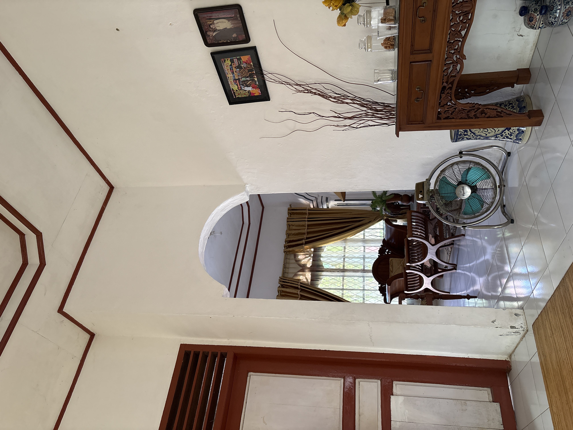 The image depicts an interior scene within a residential property in Uleepulo, Indonesia. The view is directed towards an archway that opens into another room. In the foreground, to the left, a portion of a red and white door is visible, with horizontal slatted panels at the top. The floor is tiled with light-colored squares.
Dominating the right side of the foreground is a vintage metal oscillating fan with a turquoise blade guard, positioned on the tiled floor. Next to the fan is an ornate wooden console table with carved detailing and two drawers. On the table, there is a clear glass, two glass jars containing snacks or cookies, and a vase with dried branches. To the right of the console table, stacked decorative ceramic items are visible.
Through the archway, the adjoining room features a window covered with gold curtains. In front of the window, a dark wooden dining set, including a table and chairs, is visible. The room beyond appears to be a dining area or living space.
The walls are painted white, and the ceiling has a white textured finish with dark red trim along the edges and corners. Two framed pictures hang on the right wall, one displaying a group of people and the other a portrait of a couple. The overall impression is of a traditional Indonesian home.