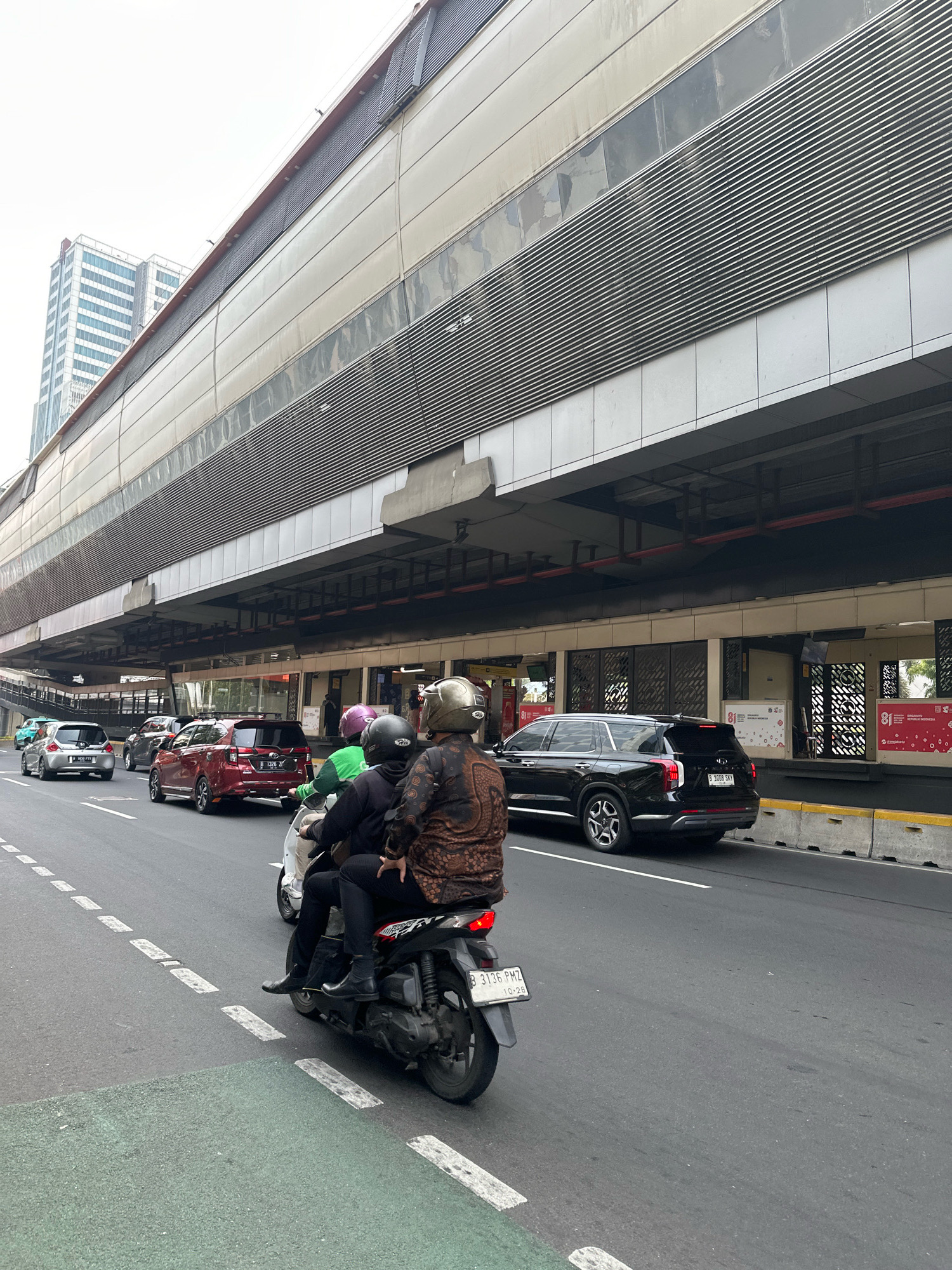 This is a medium shot, street-level, eye-level, full color, outdoor photograph taken from the perspective of a vehicle traveling on a multi-lane road. The scene depicts a city street in South Jakarta, Indonesia, dominated by a large, elevated structure that appears to be a modern building or transportation hub. This structure has a light-colored, textured facade with horizontal slat detailing and several glass panels. Below this structure, a sidewalk or pedestrian walkway is visible, featuring several shop entrances and advertisements.
In the foreground, a motorcycle with three occupants is traveling on the road. The driver and two passengers are wearing helmets. The driver is wearing a patterned brown jacket and dark pants, and the rear passenger is also wearing dark pants and a helmet. The middle passenger is wearing a green top and a black helmet. The motorcycle has a license plate with the registration "B 3136 PMZ".
Several cars are also present on the road. Closest to the motorcycle is a dark-colored SUV, possibly a Kia Telluride, with a license plate "B 1008 SKY". To the left of the SUV, a red SUV is visible, with a license plate "B 1326 ZT". Further down the road, a silver hatchback and other vehicles are present, indicating moderate traffic. The road is asphalt with visible lane markings and a green-painted bike lane.
The background includes two tall, modern buildings, suggesting a commercial or business district. The sky is overcast and bright white, typical of an urban environment. The overall impression is a bustling street scene in a contemporary Indonesian city.