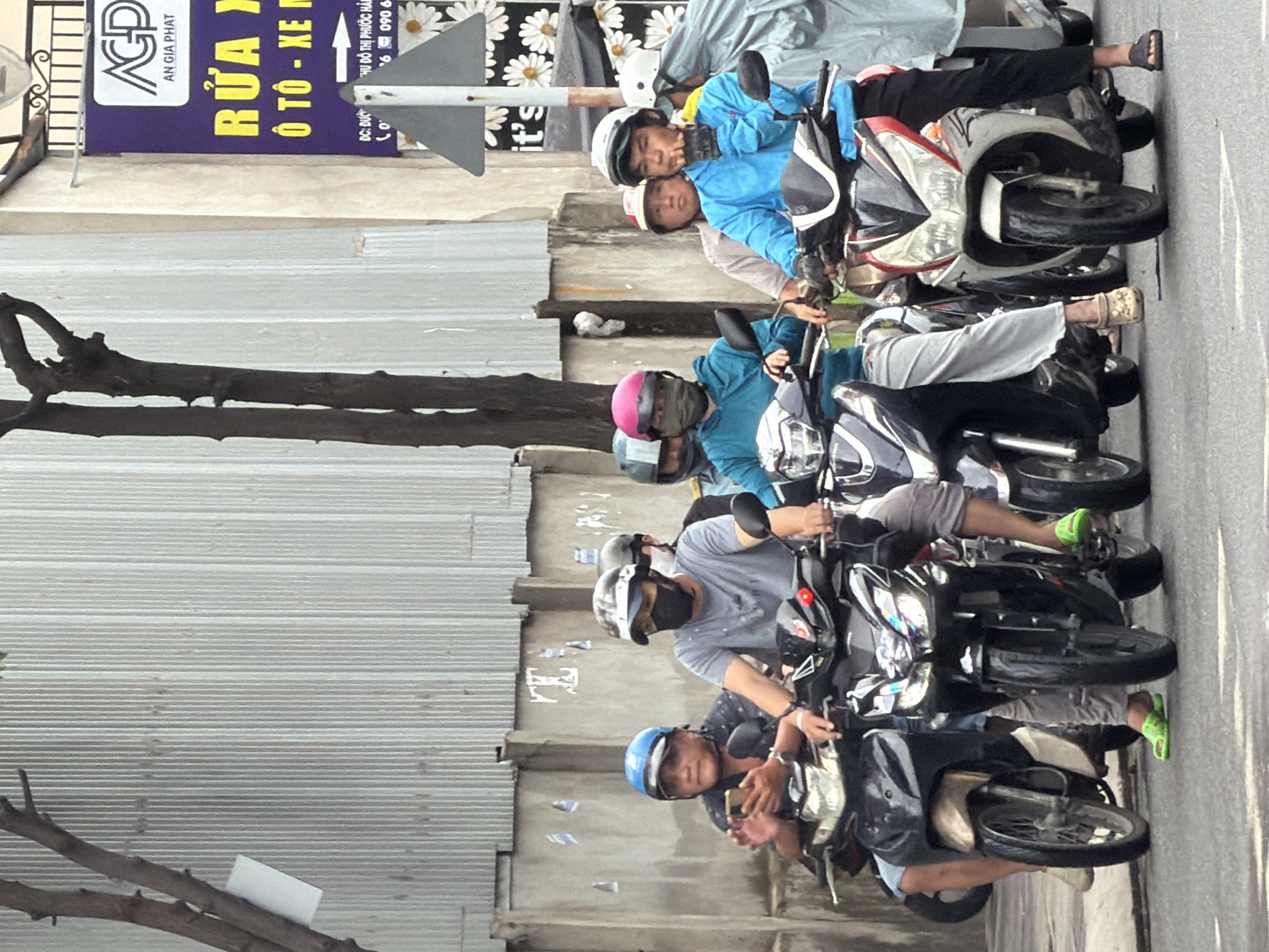 Several people are riding motorcycles in what appears to be a city street in Nha Trang, Vietnam. The weather is overcast. On the left, a man wearing a blue helmet is looking at his smartphone while riding a motorcycle. Next to him, another man wearing a grey shirt and a black face mask is riding a motorcycle. In the background, a group of people on motorcycles are also visible. The street is paved, and there are buildings and a corrugated metal wall in the background. A sign with Vietnamese text is visible on a building. Some of the people are wearing helmets and face masks, likely for protection from the elements or pollution. The overall scene depicts everyday urban life with a focus on motorcycle transportation.