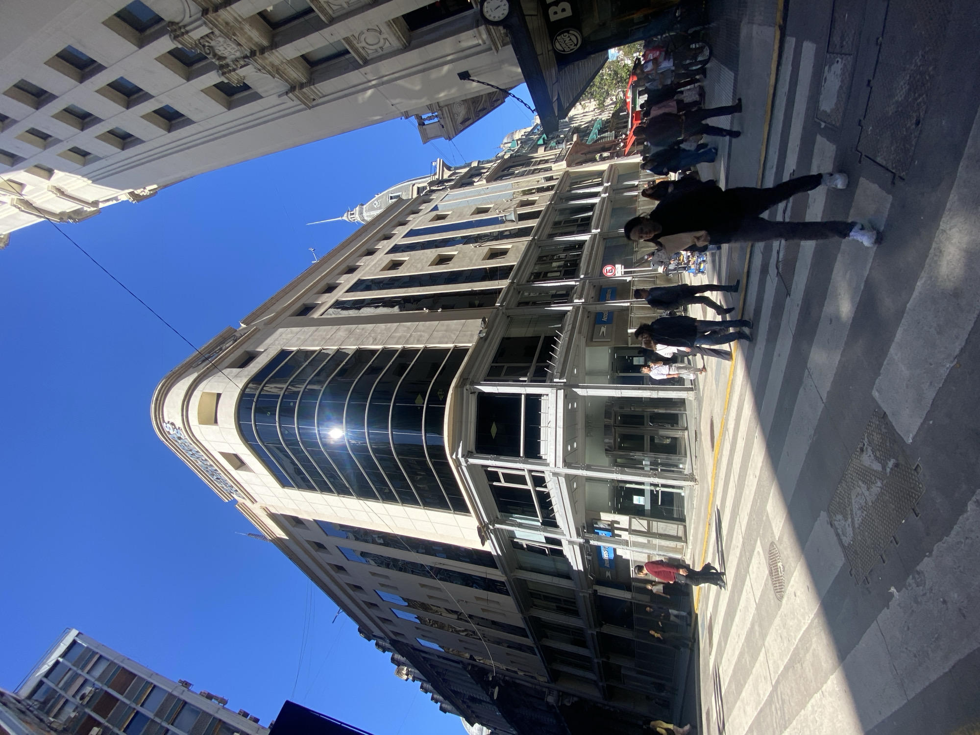 The image, taken with a vertical orientation and then rotated, depicts a sunlit urban street scene in Buenos Aires, Argentina, under a clear, bright blue sky. Tall buildings line the street, creating a dynamic perspective.
A prominent modern building with a curved corner, light-colored facade, and extensive dark glass windows dominates the right side of the frame. Sunlight intensely reflects off these windows, suggesting a bright day, likely midday or early afternoon. On the opposite side, an older building with ornate architectural details and square windows is partially visible. Further down the street, another building with a distinctive spire-like top is silhouetted against the sky.
Numerous pedestrians are visible on the street and sidewalks, indicating a busy urban environment. In the mid-ground, a person wearing a dark jacket, dark pants, and white sneakers walks across a zebra crossing, holding a light-colored object. Other people are dispersed, either walking or waiting.
Notable details include a red circular "No Parking" sign (with a crossed-out "E" for "Estacionamiento") on a pole near the modern building. Below it, a blue sign with white text partially reads "Aeropuertos". A hanging sign from an upper building shows a clock and portions of the "BURGER KING" logo and text, confirming a commercial area. The street features a clearly marked zebra crossing with white and gray stripes, and a yellow curb line. Manhole covers are visible on the asphalt.