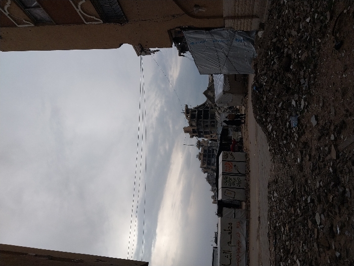 The image captures a devastated urban environment in Khan Yunis, Palestinian Territory, under an overcast sky. In the foreground, a large mound of dark rubble, consisting of fragmented concrete and debris, occupies the lower center. The ground is unpaved and strewn with additional refuse. The midground features a series of makeshift shelters or tents, constructed from tarpaulins and various materials. One shelter on the left prominently displays partial Arabic text, "مطعم الشا" (possibly part of "Al-Shawe Restaurant"). Another shelter exhibits handwritten messages or graffiti. Two individuals, one in dark clothing and another in lighter attire, are observed standing or walking between these shelters. Overhead, multiple utility lines traverse horizontally across the frame. In the background, several multi-story buildings appear severely damaged, with large sections missing, exposed rebar, and compromised facades. Portions of damaged buildings frame the scene on the left and right, with the structure on the right showing visible cracks and decorative arch-like elements above windows.