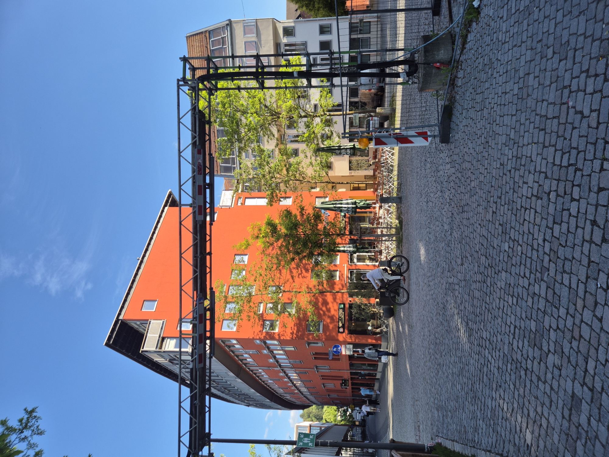 In the city of Landshut, Germany, a street scene unfolds under a clear blue sky, with the sun casting long shadows. The foreground is paved with cobblestones, leading towards a curved, vibrant orange building that dominates the left and center of the frame. This modern building features numerous windows and a distinctive slanted roofline. To the right of the orange building, a lighter-colored building with a pitched roof is visible.
In the middle ground, a person is riding a bicycle. Several other pedestrians are walking along the sidewalk in front of the orange building. Trees with bright green foliage are interspersed between the buildings, adding natural elements to the urban landscape. Outdoor seating areas with chairs and tables are set up in front of some establishments, suggesting a commercial or dining area.
A prominent metal structure, resembling scaffolding or a temporary overhead support system, crosses the image from left to right, partially obscuring the buildings and sky. A construction barrier with red and white striped markings and a concrete base is visible on the right side of the scene, indicating ongoing work. The overall impression is a dynamic urban environment with a mix of modern architecture and street life.