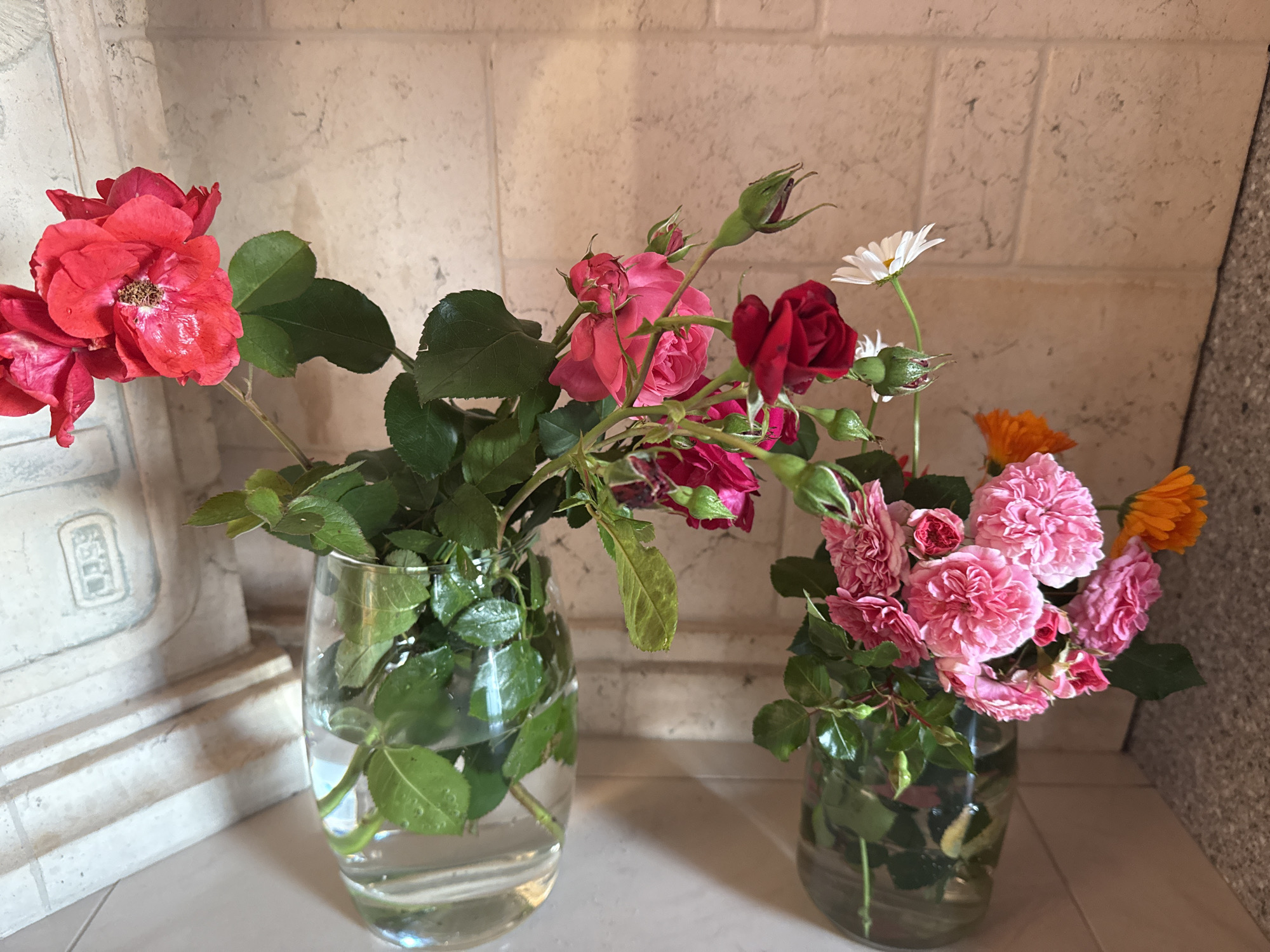 The image shows two glass vases filled with water and flowers, placed on a white surface against a textured beige and grey wall. The vase on the left contains a bouquet of red roses with green leaves. The vase on the right holds a mix of pink roses, a white daisy, and orange marigolds, also with green leaves. The lighting suggests an indoor setting, likely during the daytime. There are no people visible in the image. The background wall appears to be made of stone or brick, possibly in a building in Saint Petersburg, Russia.