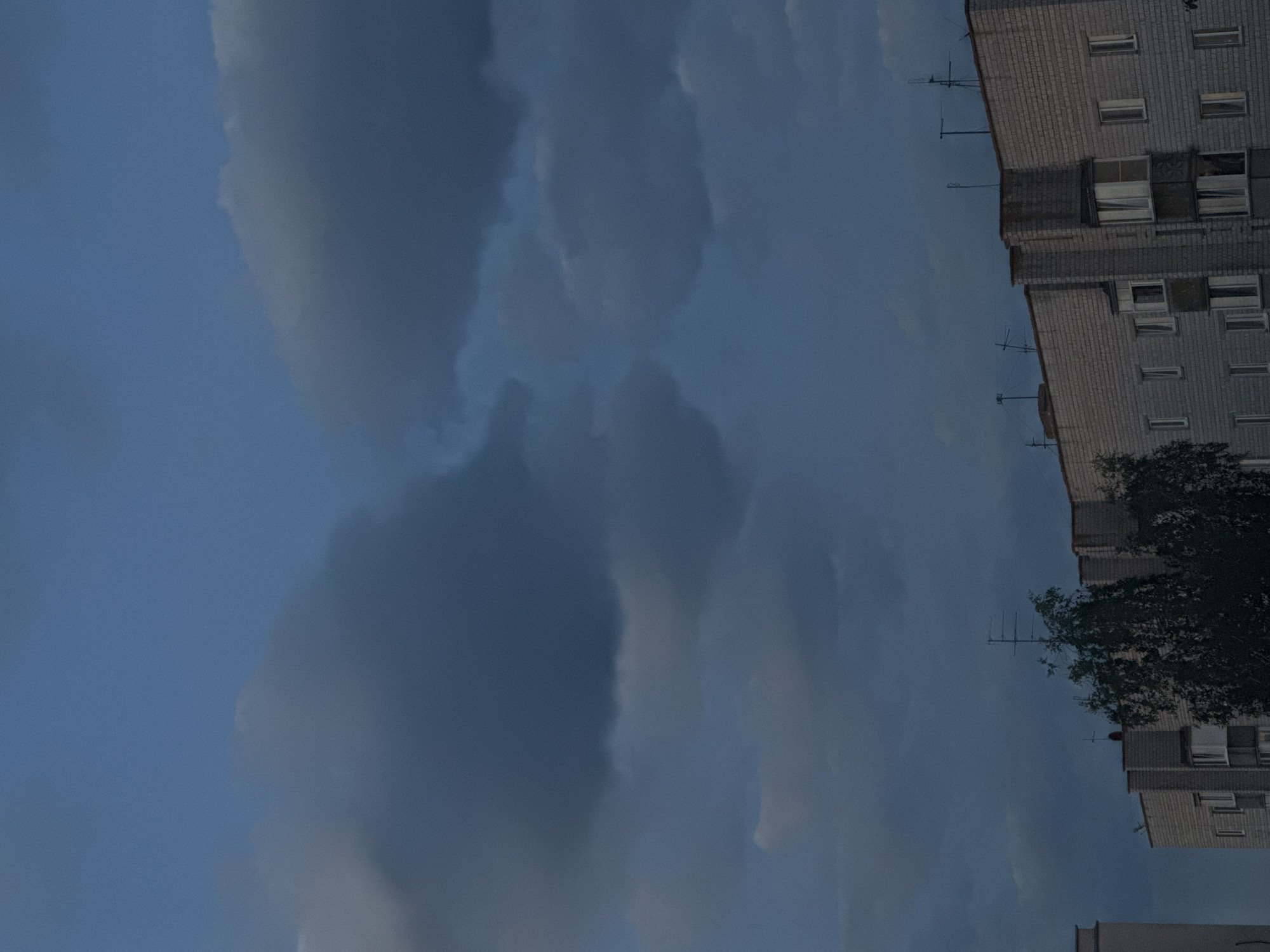 The image shows a view of apartment buildings against a darkening, cloudy sky in the Yarominski Sel'ski Sovet, Belarus. The buildings are constructed of light-colored brick and appear to be multi-story residential structures. Several antennas are visible on the rooftops. A tree with dense foliage is positioned in the foreground, partially obscuring one of the buildings. The sky is filled with heavy, dark clouds, suggesting an overcast or approaching storm. There are no people visible in the image.