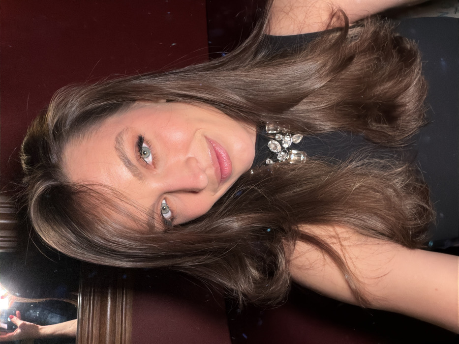 A woman with long, wavy brown hair and green eyes smiles at the camera. She is wearing a black dress with a jeweled collar. The background is dark red and there is a reflection of her holding a phone in her left hand in a mirror. The image is slightly tilted.