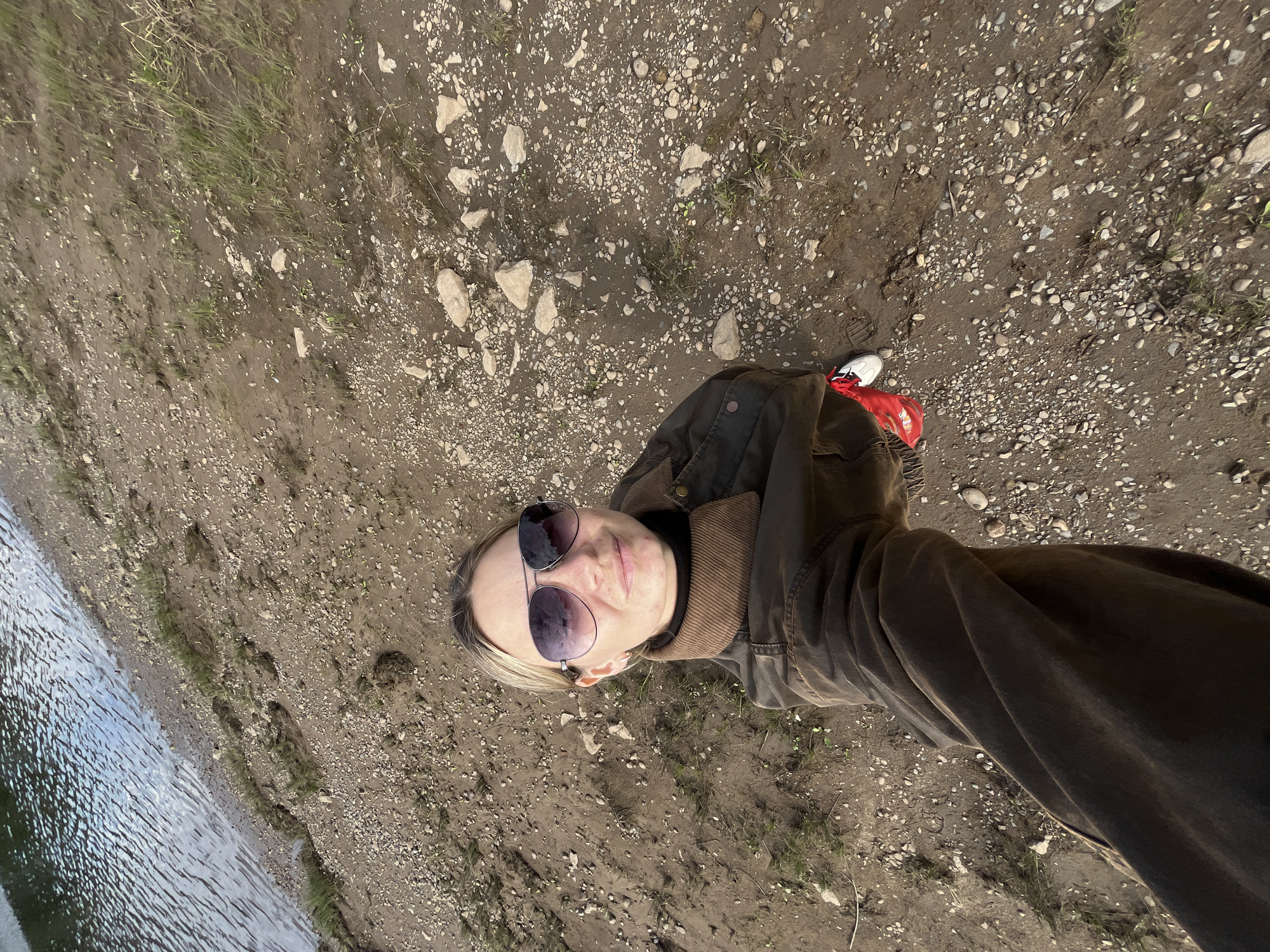 The image shows a selfie taken from a high angle, looking down at a person standing on a muddy, rocky shore next to a body of water. The person is wearing sunglasses, a brown jacket with a corduroy collar, and white sneakers with red accents. The background shows a rippling body of water with reflections of the sky. The ground is a mix of dark mud, small stones, and sparse patches of grass. The scene is consistent with a natural outdoor environment, possibly a riverbank or lake shore. The specific location in Chaikovsky, Russia, is not discernible from the visual information provided in the image.