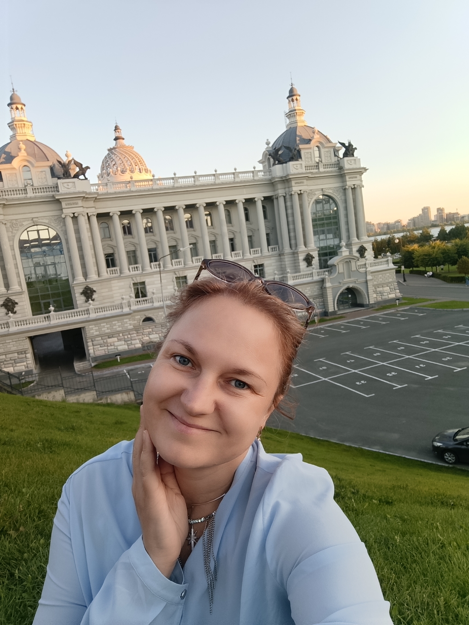 A woman with light brown hair and blue eyes smiles at the camera, holding her hand to her chin. She is wearing a light blue collared shirt and a silver necklace with a cross pendant. Sunglasses are perched on her head. In the background, a grand white building with classical architecture, including columns and domes, is visible. The building appears to be the Ministry of Agriculture building in Kazan, Russia, characterized by its distinctive onion domes and ornate facade. To the right of the building is a large, paved parking lot with marked parking spaces. A grassy slope is in the foreground, and a black car is parked in the background. The sky is clear and light blue, suggesting it is daytime.