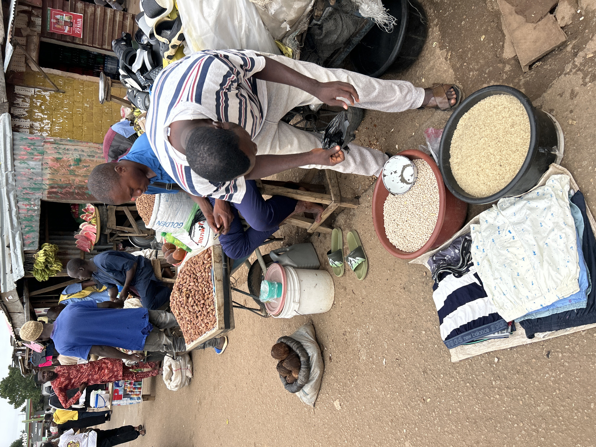 The image depicts a busy street market in Jos, Nigeria. A man in a blue shirt and a patterned cap stands facing away from the camera, near a stall with a large display of peanuts. Beside him, another man in a striped t-shirt and light-colored pants is bent over, holding what appears to be a dark plastic bag. In the foreground, several items are laid out on the ground, including folded clothing, a large tub of rice, and a smaller bowl of white beans. A sack filled with coconuts is also visible. In the background, other market stalls are set up, with vendors and shoppers interacting. The stalls feature various goods, including fruits like bananas and watermelon slices, and displays of shoes and other items. The environment appears to be outdoors, with a paved ground and corrugated metal roofs over the stalls. The overall scene conveys the hustle and bustle of a typical Nigerian market.
