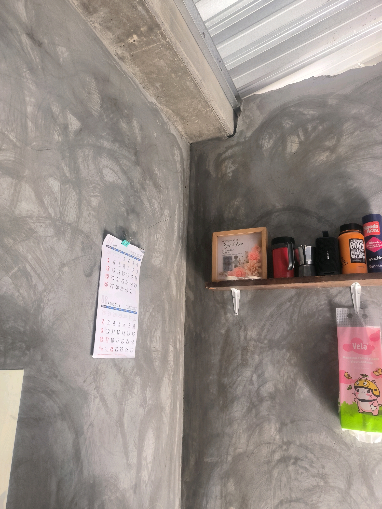 A calendar for July and August is hanging on a textured gray wall in what appears to be an unfinished or industrial-style room. The calendar is partially obscured by a blue clip. To the right, a wooden shelf is mounted on the wall, holding several items: a framed picture with pink flowers, a red thermos, a metal coffee maker, a black portable speaker, an orange jar labeled "EVERYBODY BORN DIFFERENT EVERYBODY BORN WELLBORN", and two blue cylindrical containers, one with red lettering. Below the shelf, a pink package of facial tissues with cartoon images of cats and butterflies is hanging. The room has a corrugated metal ceiling and concrete beams. The overall impression is of a utilitarian space with personal touches.