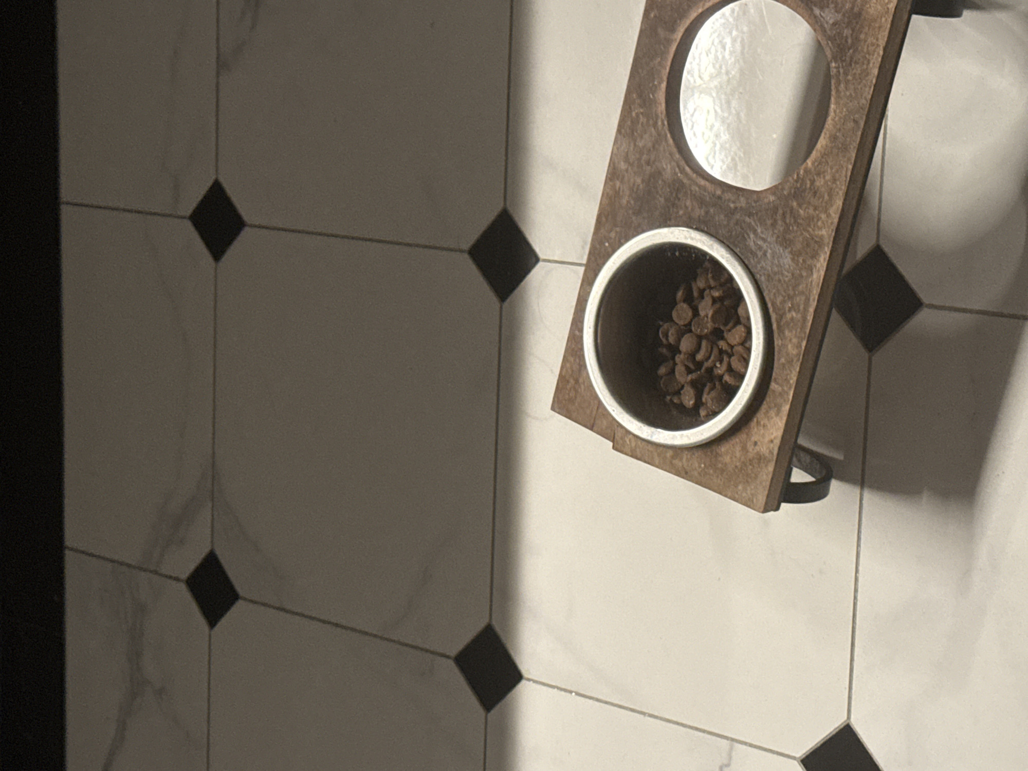 In an indoor setting, the image shows a close-up view of a raised wooden dog bowl stand. The stand has two circular cutouts, one of which contains a metal bowl filled with brown kibble. The stand is positioned on a tiled floor composed of large white tiles with marbled grey veining, interspersed with smaller black diamond-shaped tiles. The lighting suggests a natural light source casting shadows across the floor and the stand. The overall impression is one of a domestic setting, likely a home. The location is unknown.